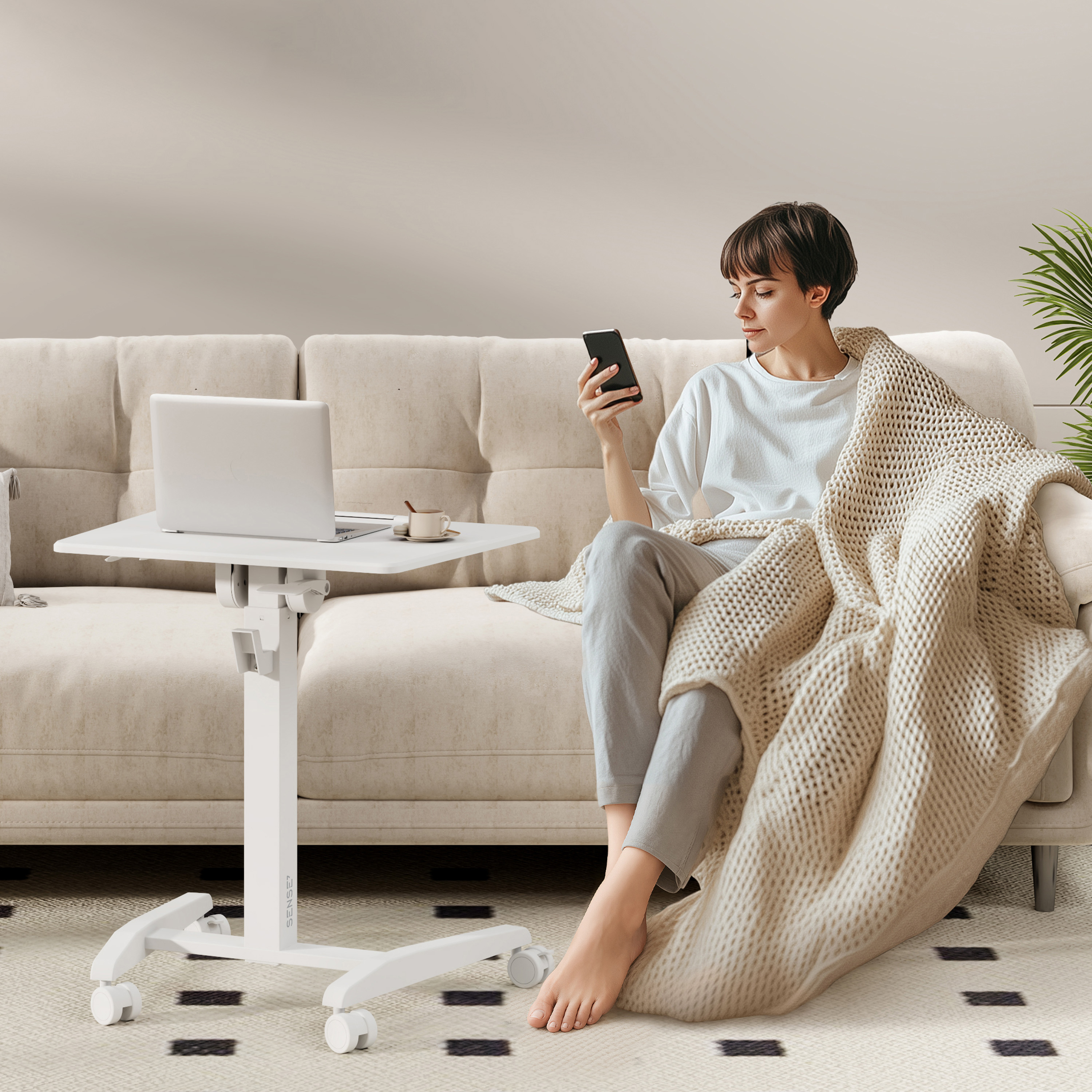 Mujer sentada en un sofá, usando un teléfono, con una laptop y un escritorio blanco ajustable. Está cubierta con una manta.