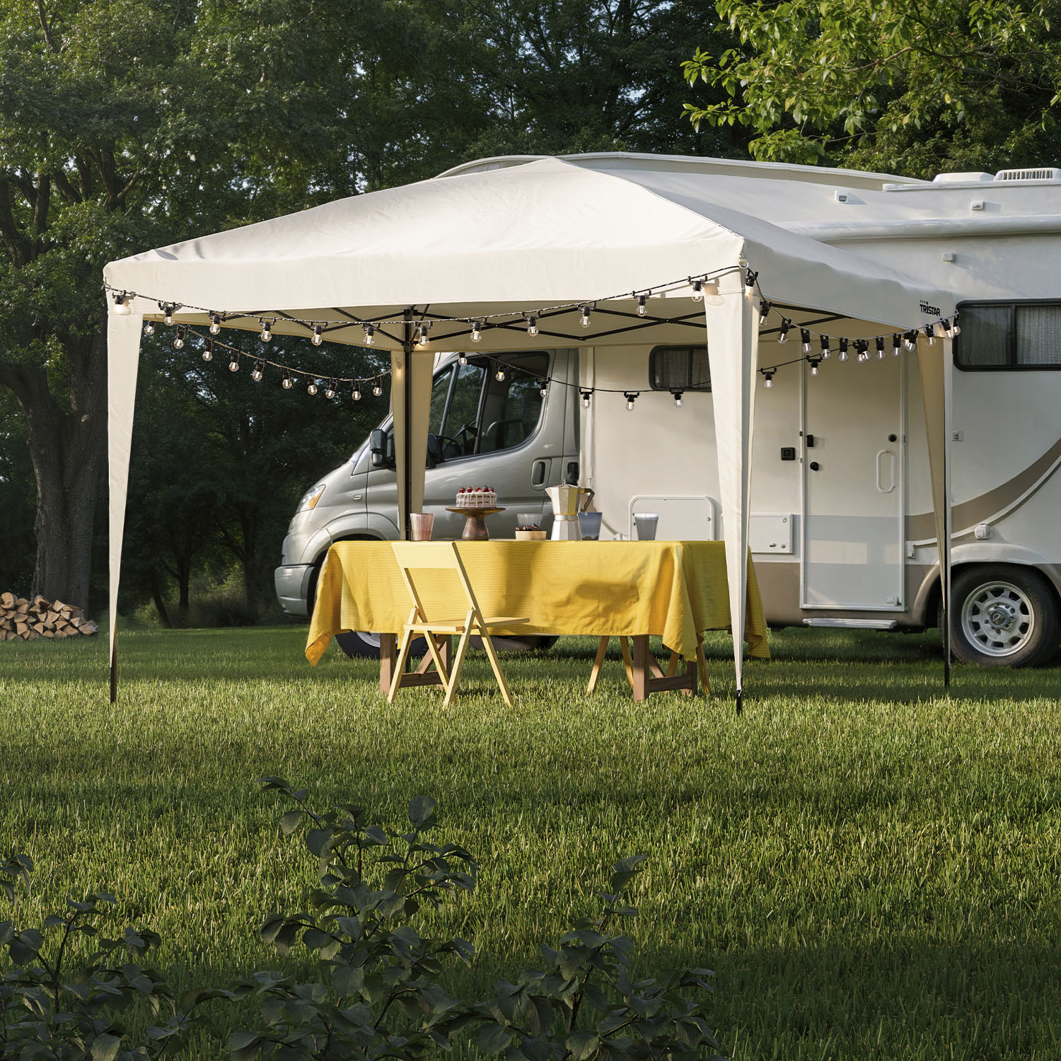 Een gazebo met tafel, stoelen en gele doek, naast een camper en lichtslingers.