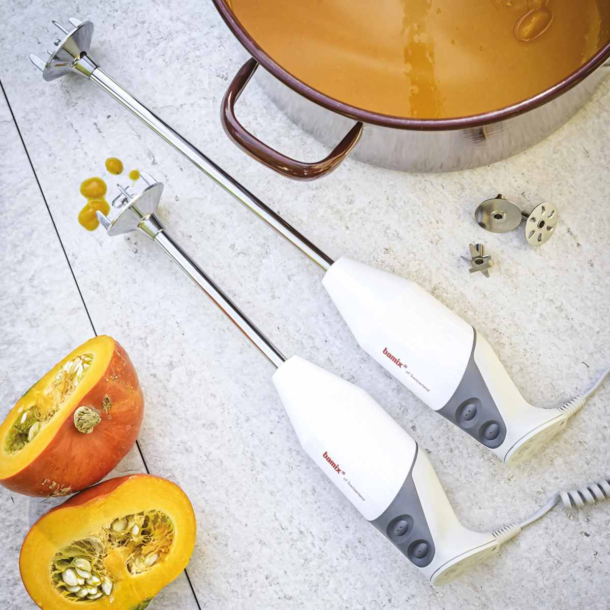 Zwei Handmixer, Kürbishälften und ein Topf Suppe. Die Mixer und der Kürbis stehen auf einem Steintisch.