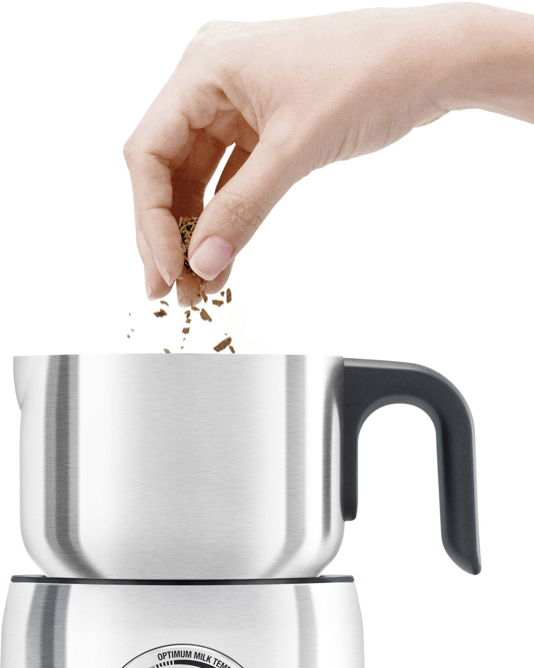 Una mano versa caffè macinato in un montalatte argentato, su sfondo bianco.