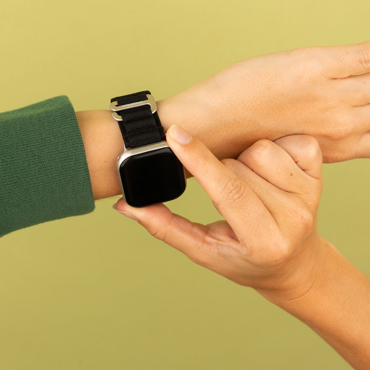 Hand van persoon die een zwarte band op een smartwatch aanpast, tegen een geelgroene achtergrond.