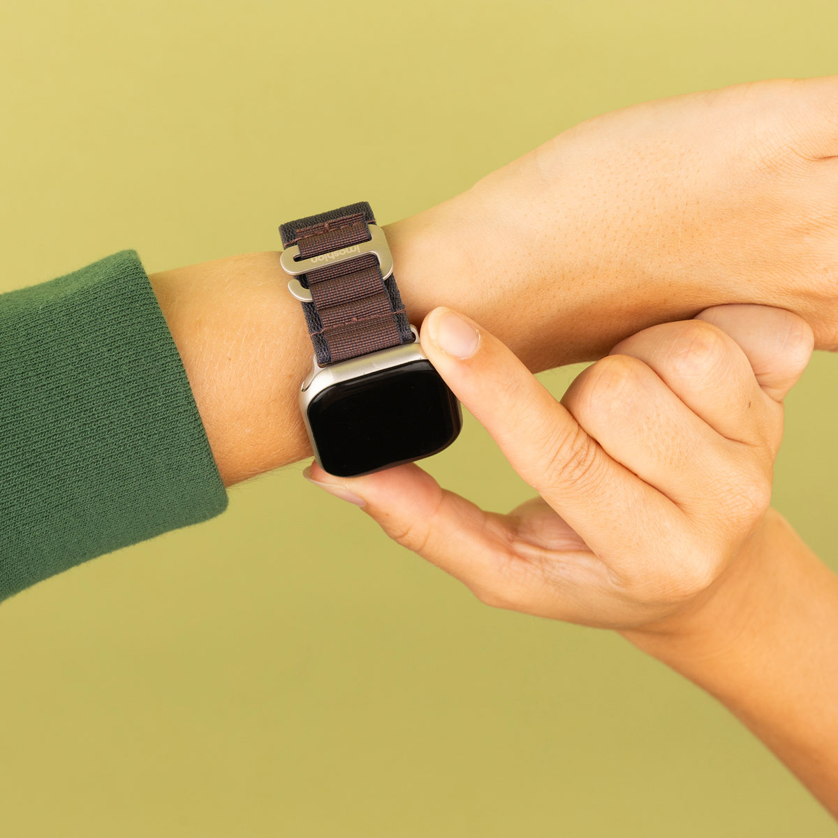 Een hand met een smartwatch met een bruin-zwarte band, op een groene achtergrond.