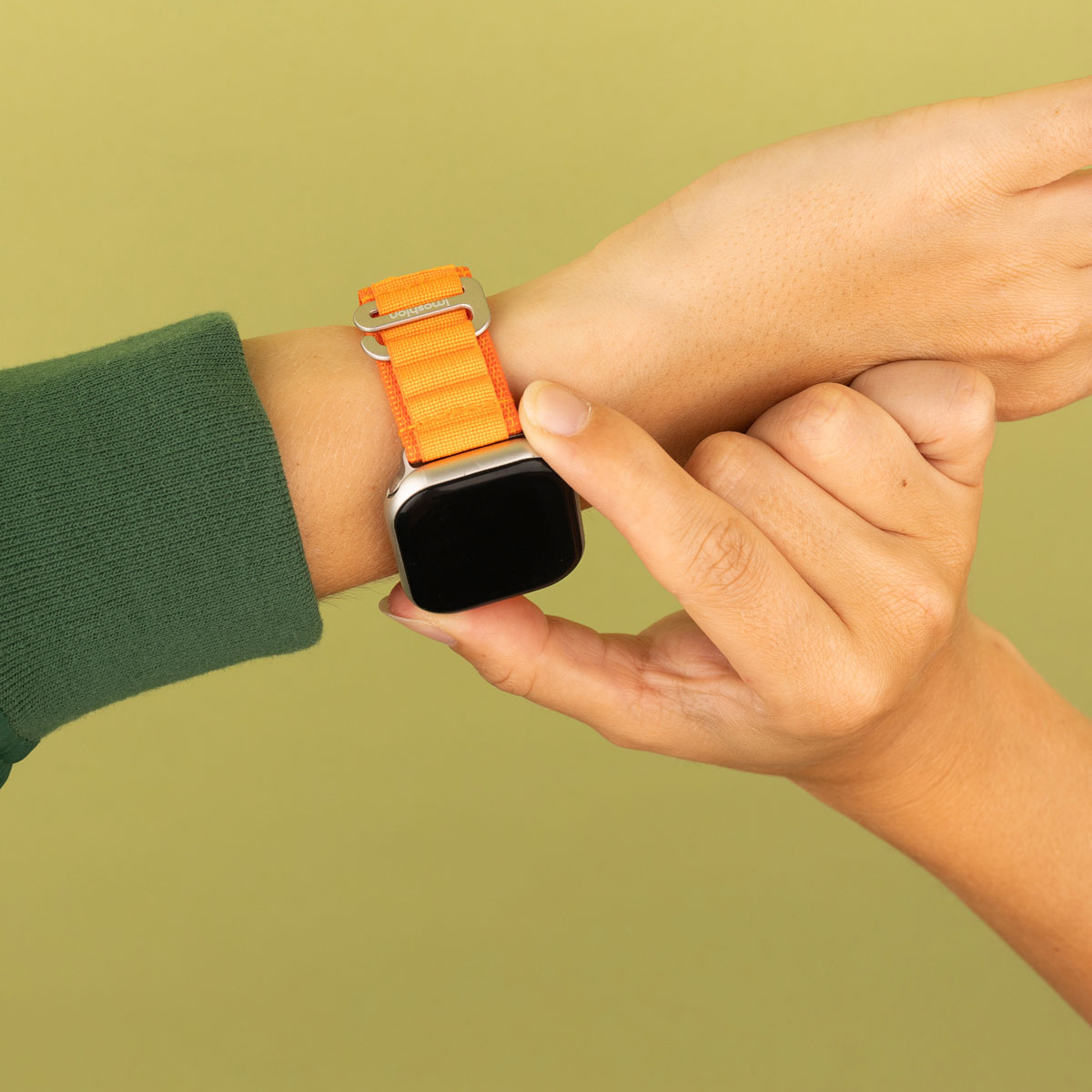 Pols met oranje horlogebandje. Een andere hand raakt de wijzerplaat aan. Groene achtergrond.