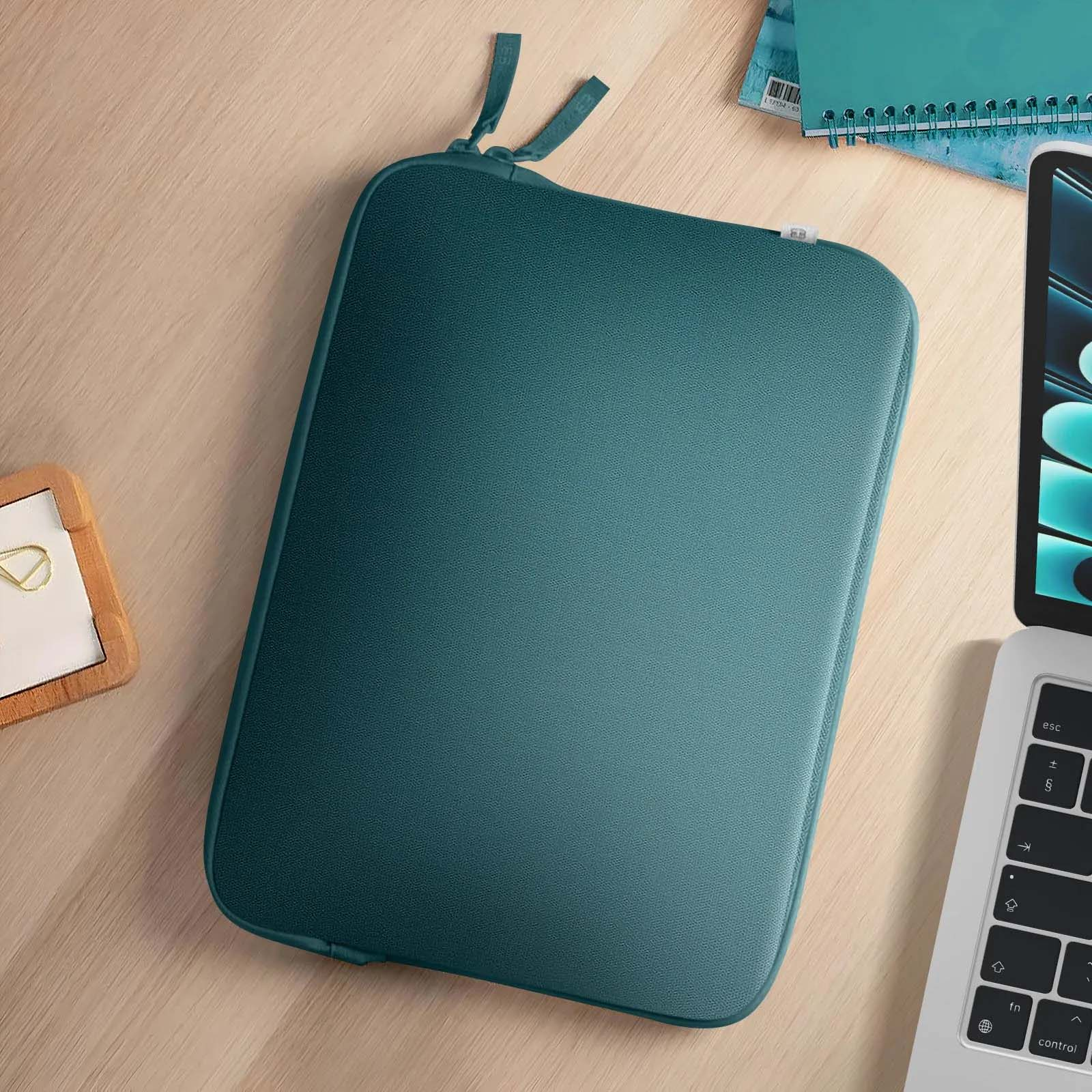 Una custodia per laptop verde acqua e accessori su un tavolo di legno. Taccuino e penna visibili.