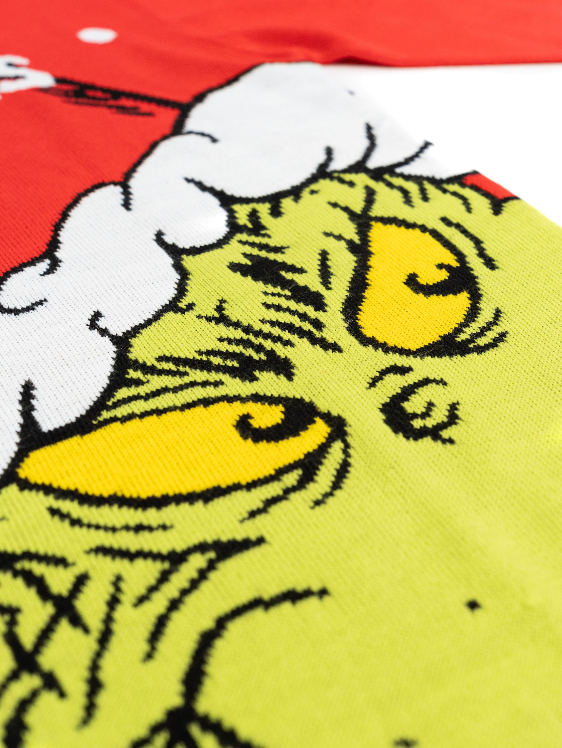 Nahaufnahme des Gesichts des Grinch auf einem roten Pullover. Zeigt seine grüne Haut, gelben Augen und weißes Fell.