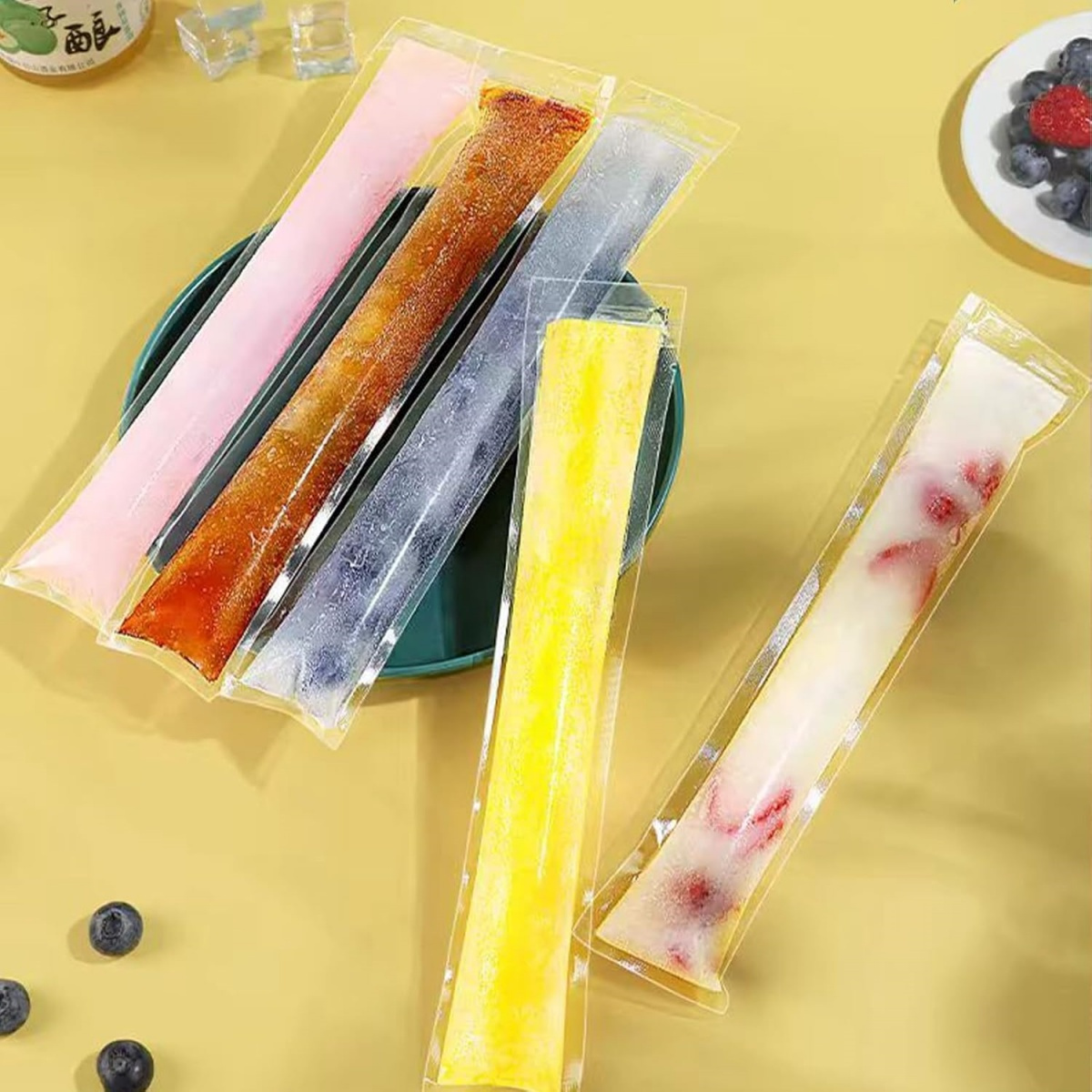 Verschiedene farbige Eis am Stiel in Plastikverpackungen, mit Blaubeeren und Erdbeeren auf einem grünen Teller.