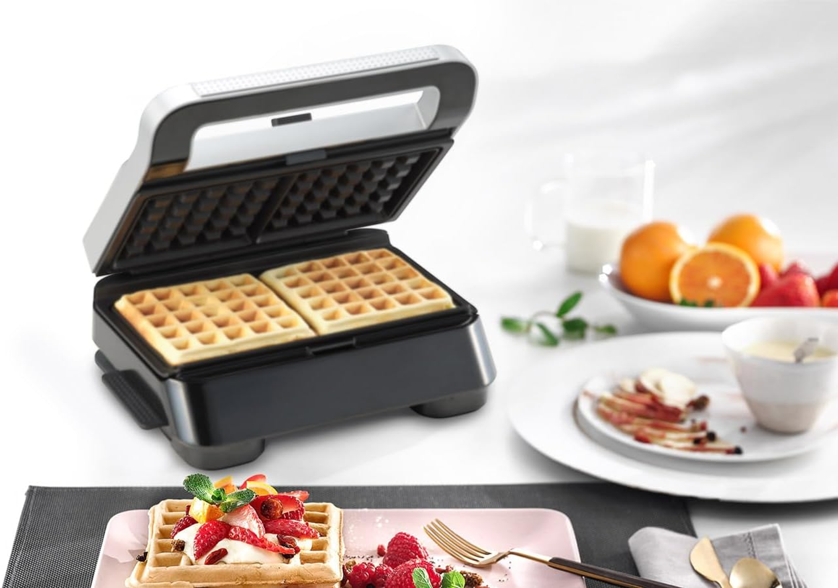 Een wafelijzer met twee wafels erin. Er staat een bord met wafels en fruit op de tafel.