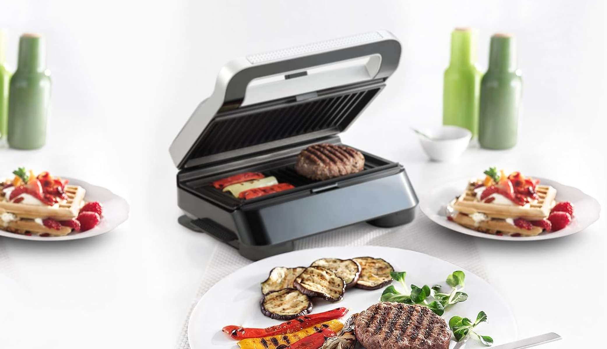 Een zwarte grill met een hamburger, rode en gele paprika en aubergine. Borden met wafels en fruit aan de zijkanten.