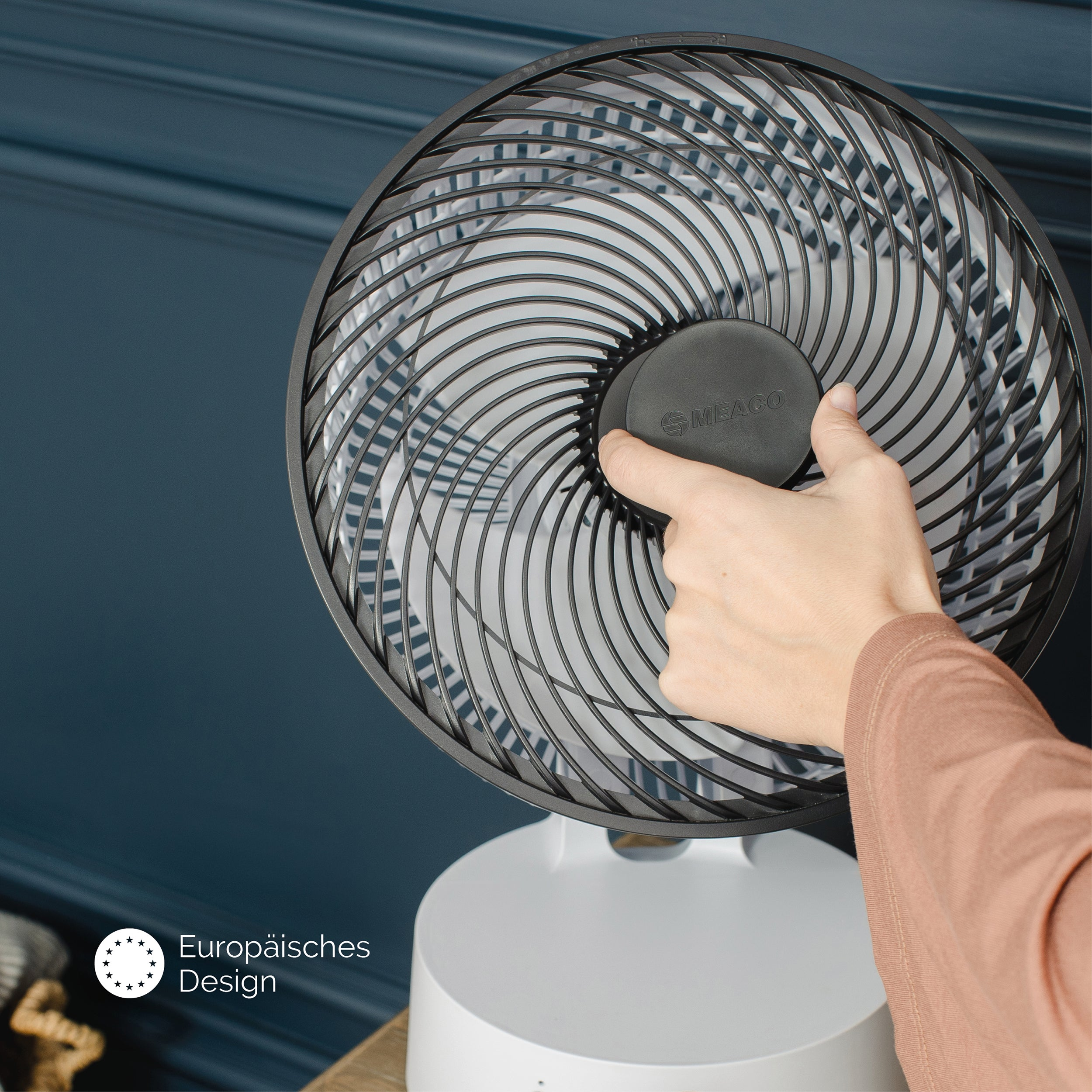 Ręka dotyka środka czarnego wentylatora. Wentylator stoi na białej podstawie. Widoczne jest logo europejskiego designu.