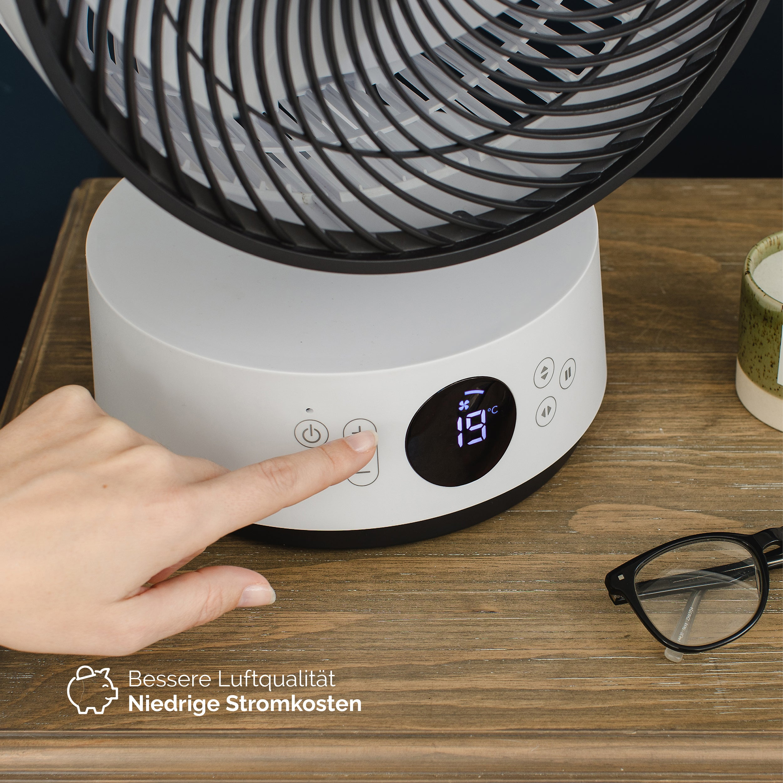 Eine Hand drückt eine Taste an einem weißen Ventilator. Der Ventilator hat eine digitale Anzeige, die die Temperatur anzeigt. Es gibt eine Brille und eine Kerze.