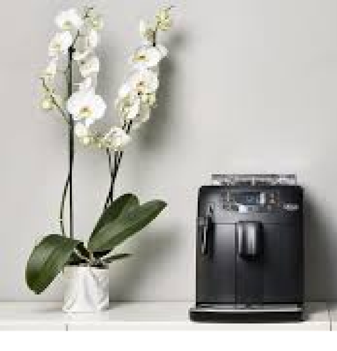 Een zwarte koffiemachine en een witte pot met witte orchideeën op een wit oppervlak tegen een grijze muur.