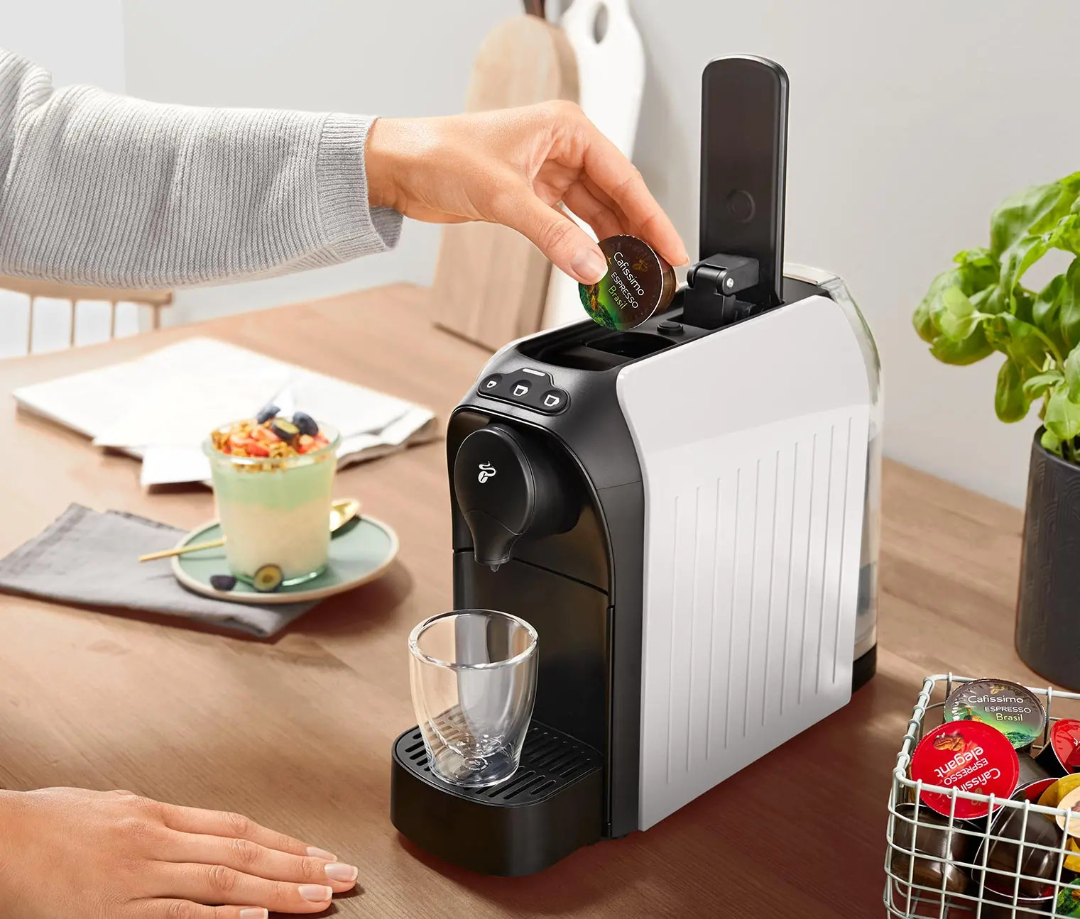 Eine Hand legt eine Kaffeekapsel in eine weiß-schwarze Kaffeemaschine ein. Ein Glas ist platziert.