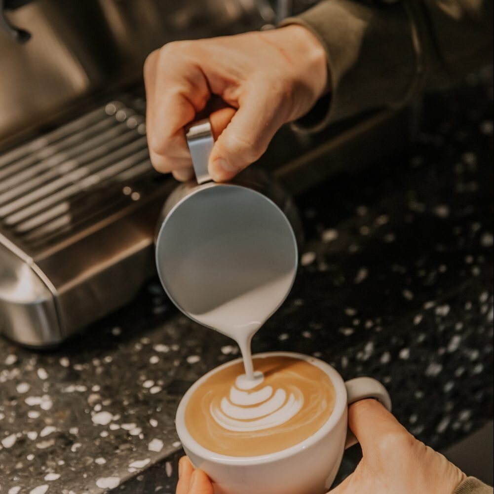 Ręce wlewają mleko z metalowego dzbanka do filiżanki kawy, tworząc latte art.