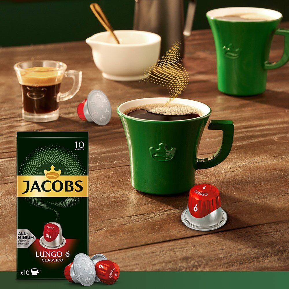 Capsule di caffè Jacobs e una tazza verde di caffè. Ci sono anche un cucchiaio e una tazzina di caffè.