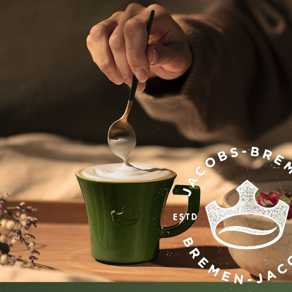 Eine Hand gießt mit einem Löffel Schaum über Kaffee in einer grünen Tasse auf einem Holztablett. Ein Logo ist sichtbar.