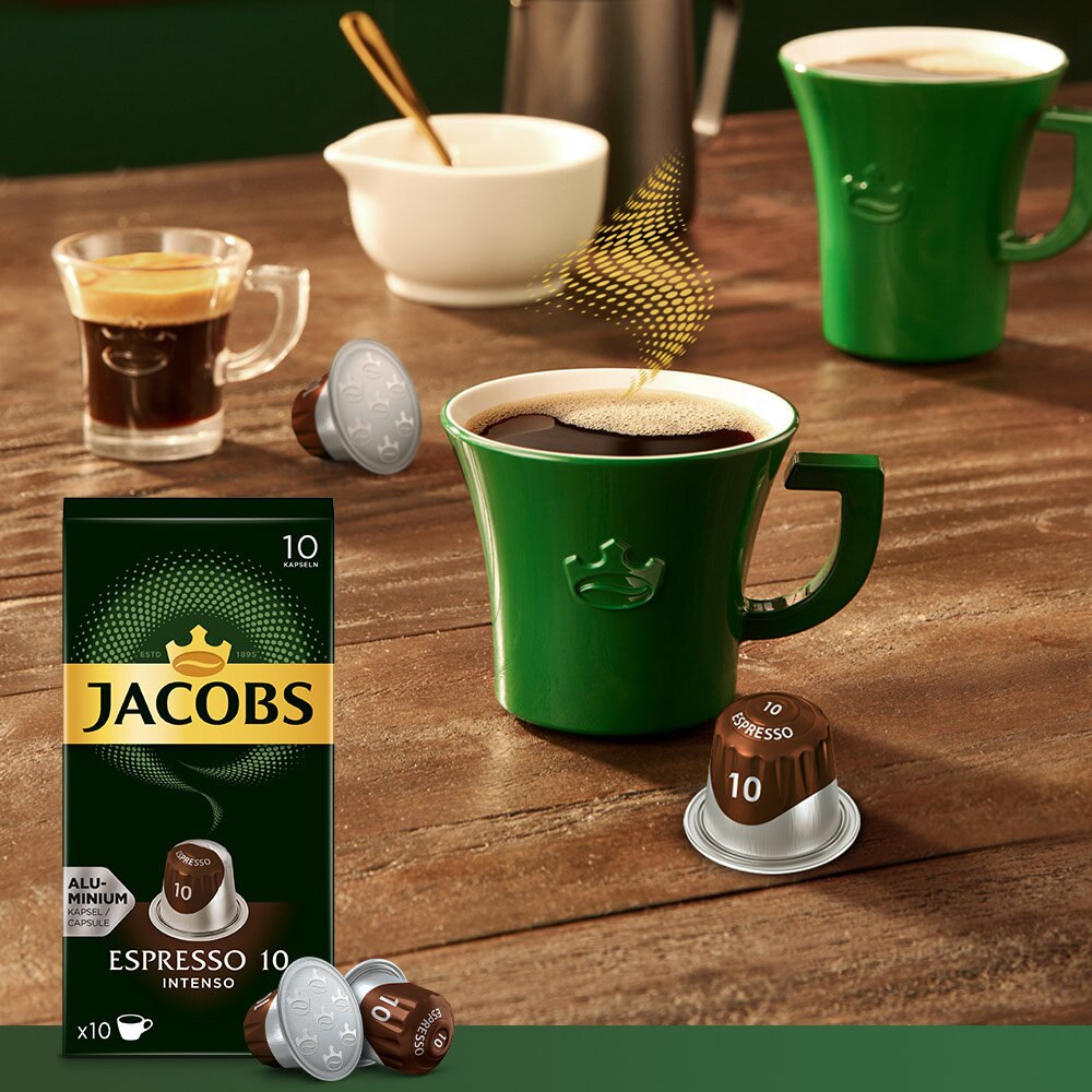 Ein Kaffee-Setup mit einem grünen Becher, einer kleinen Espressotasse, Kapseln und einer Kaffeebox auf einem Holztisch.