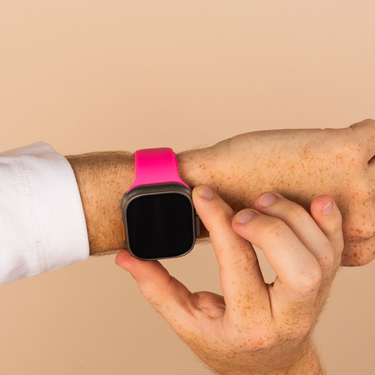 Wrist with an Apple Watch. Watch has a pink band, black screen, and silver case. Beige background.