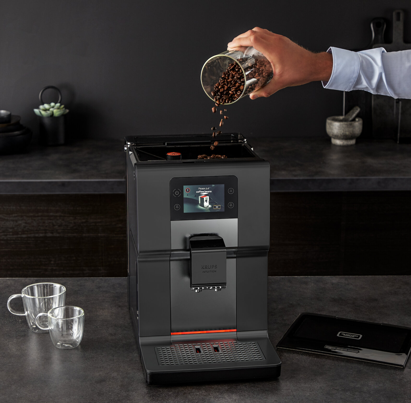 Kaffeemaschine, in die von Hand Kaffeebohnen gegossen werden, mit Tassen auf der Theke.