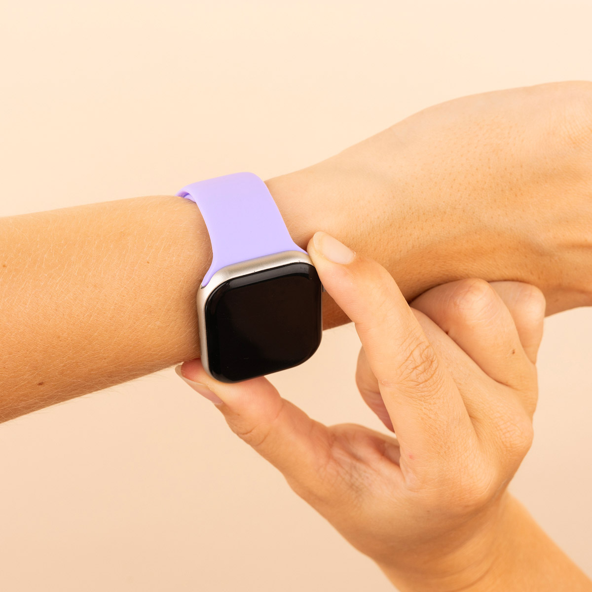 A wrist with a lavender smartwatch. A hand touches the watch face, the background is plain.