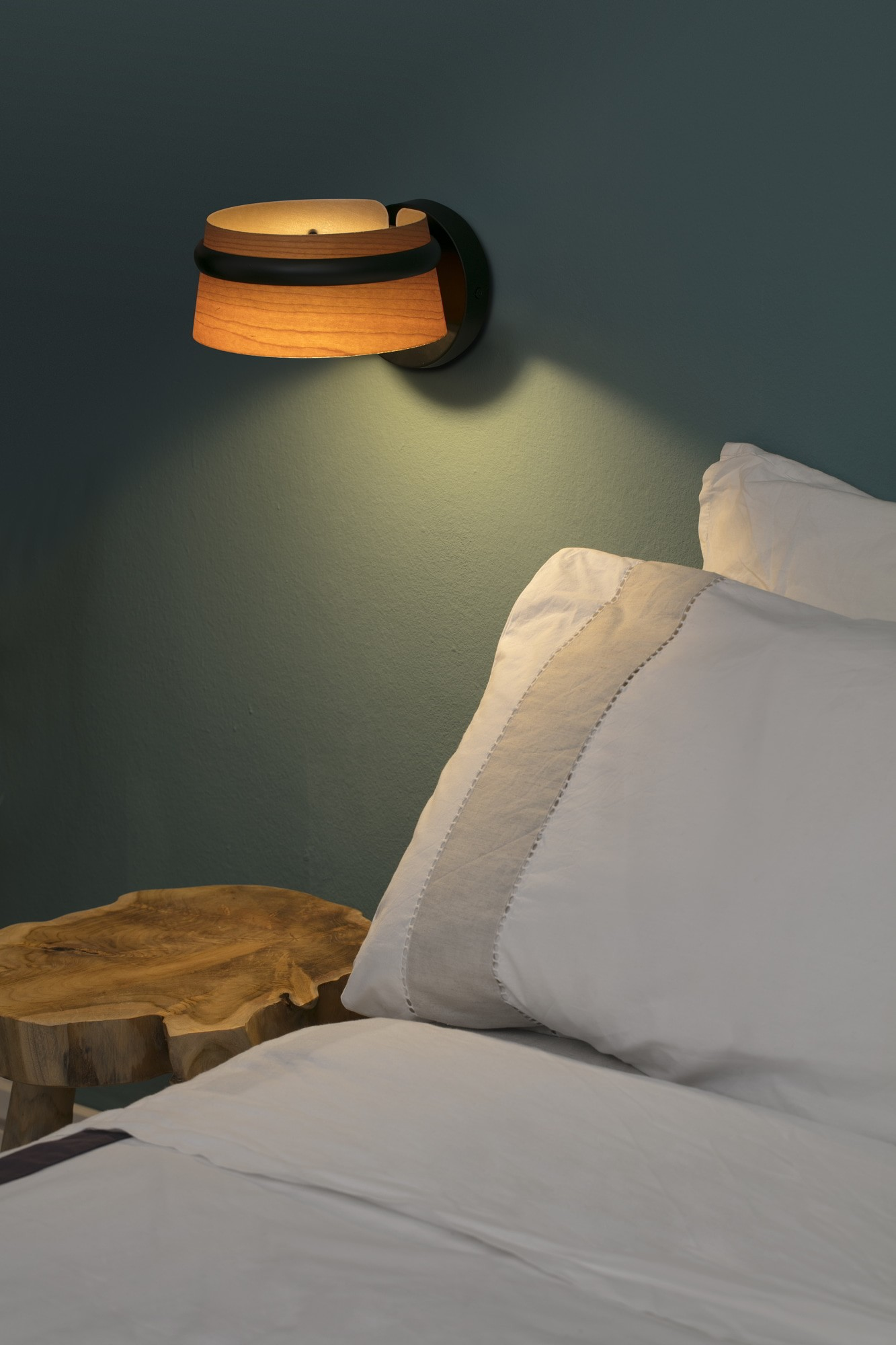Una lampada da parete con paralume in legno è accesa sopra un letto. Un comodino in legno è vicino al letto.