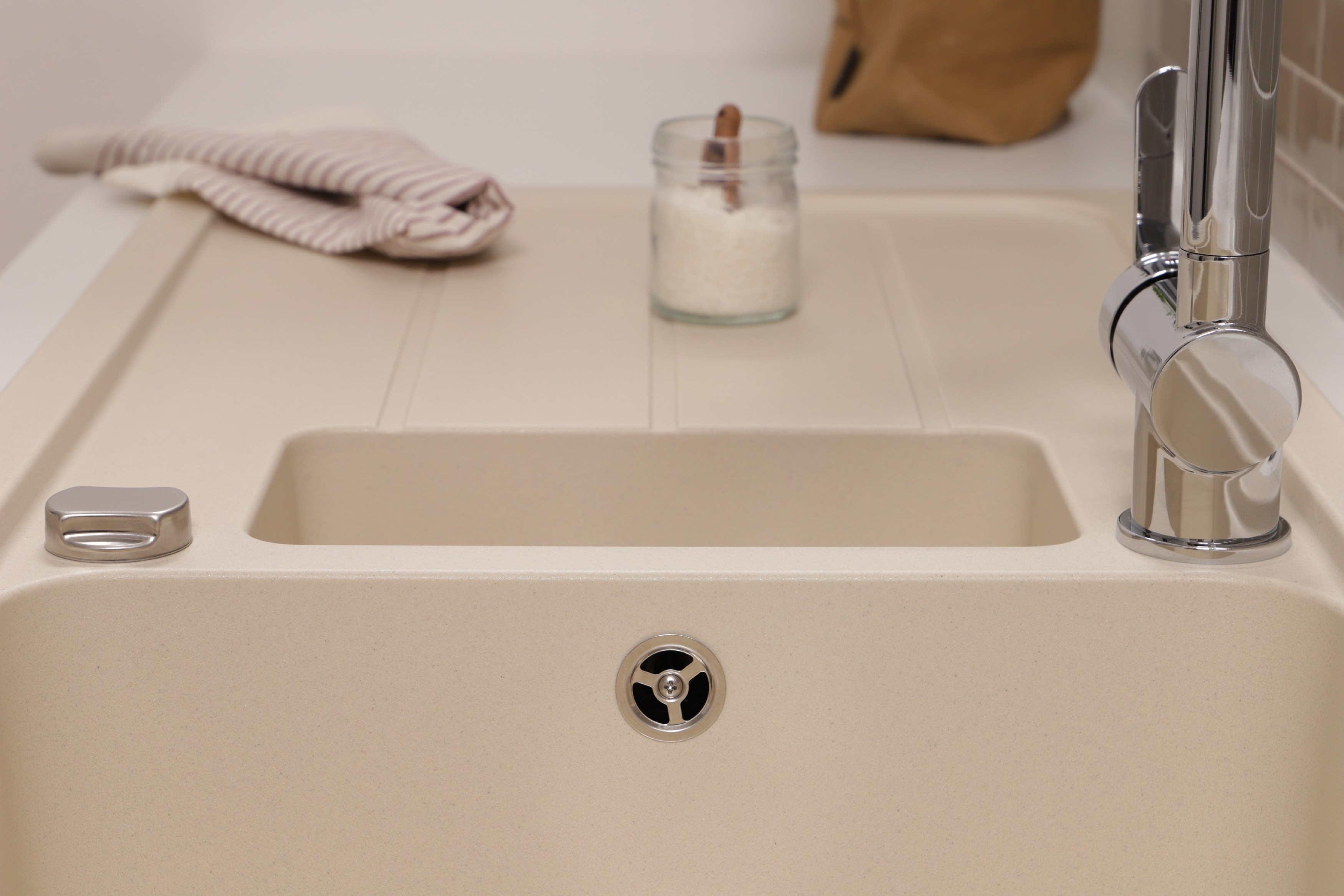 Beiges Waschbecken mit Wasserhahn, einem Glas mit weißem Pulver, einem gefalteten Handtuch und einem Abfluss. Der Hintergrund ist unscharf.