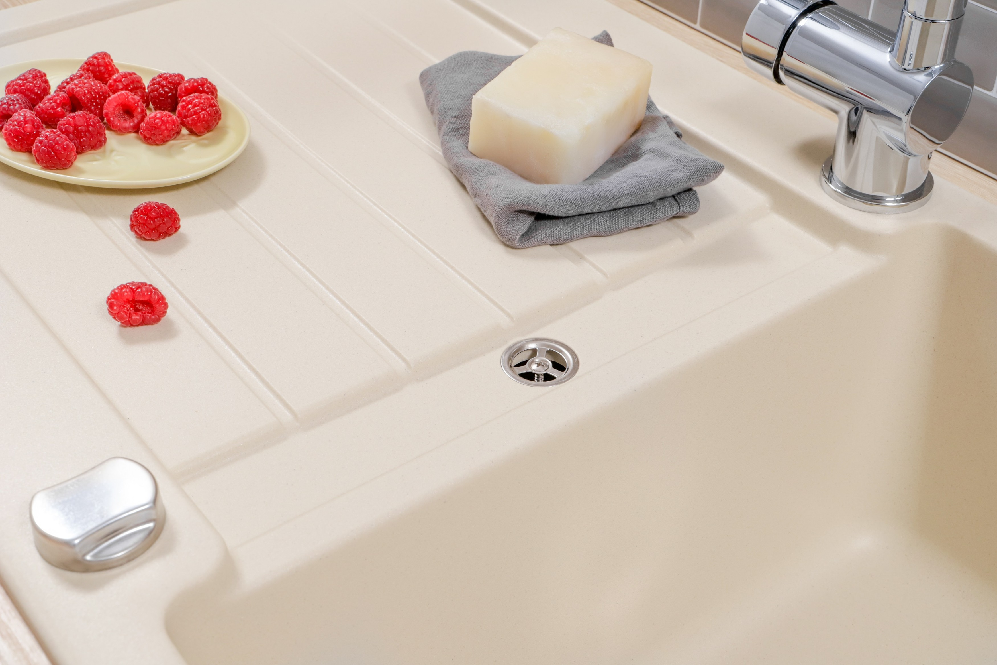 Beiges Waschbecken mit Himbeeren auf einem Teller, einem Stück Seife auf einem Handtuch und einem Wasserhahn. Der Hintergrund ist unscharf.
