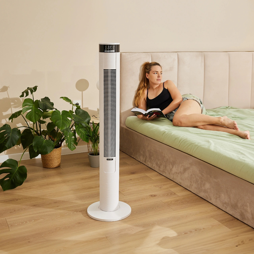 Kobieta czyta na łóżku, w pobliżu biały wentylator i rośliny. Scena jest oświetlona naturalnym światłem.