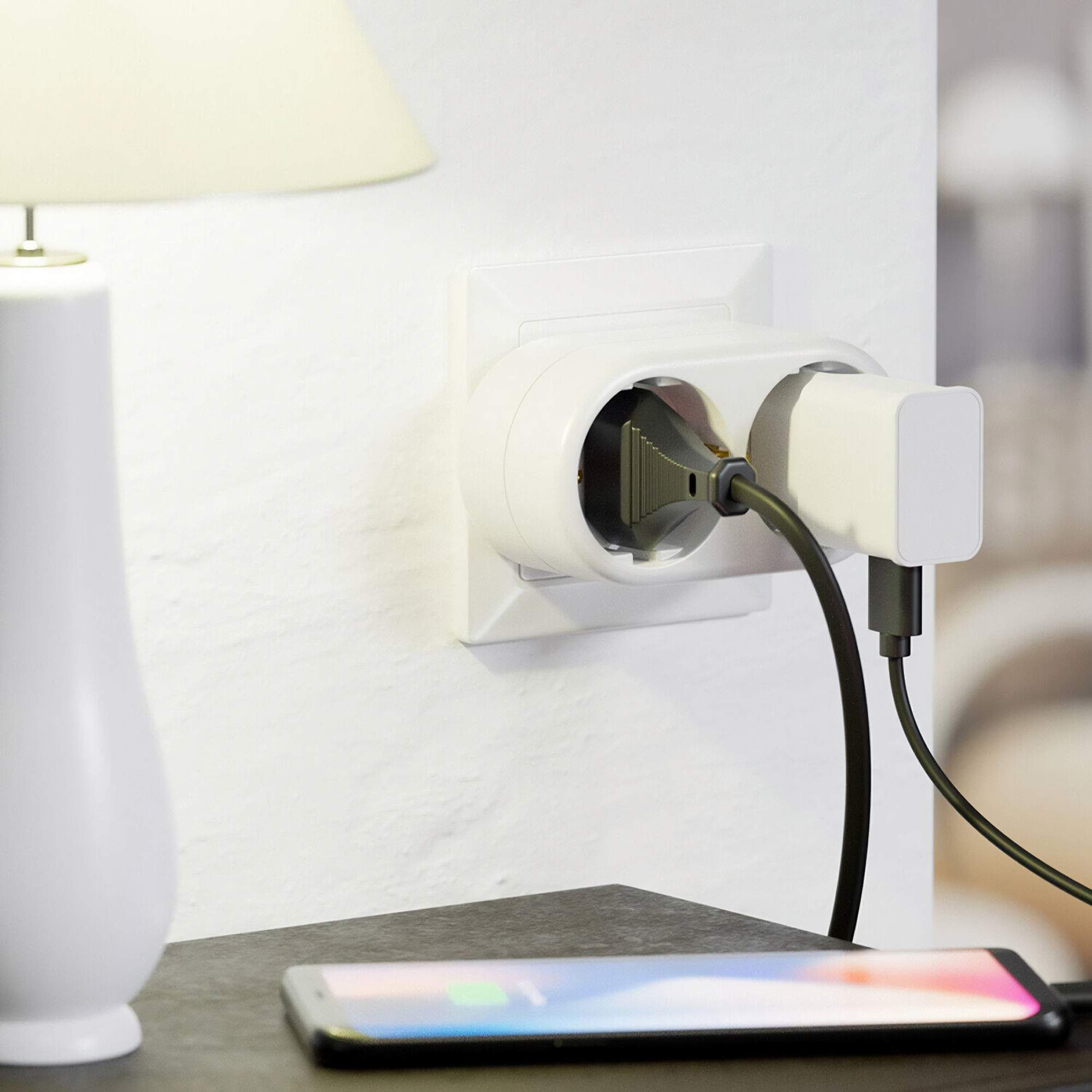 Weiße Steckdose mit eingestecktem schwarzen Kabel und weißem Ladegerät. Ein Telefon mit Ladeanzeige darunter.