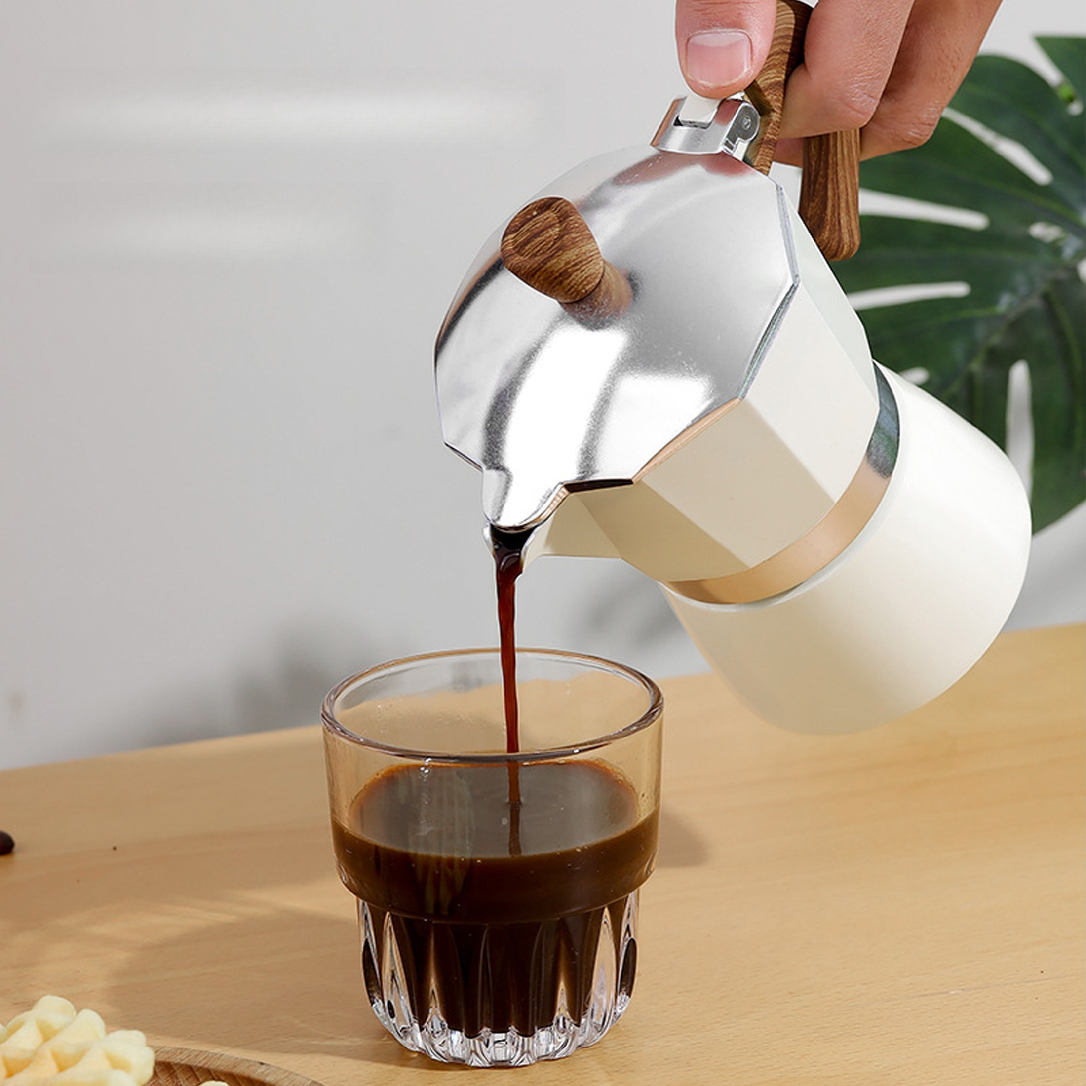 Eine Person gießt Kaffee aus einem weißen und silbernen Espressokocher in ein klares Glas auf einer Holzoberfläche.