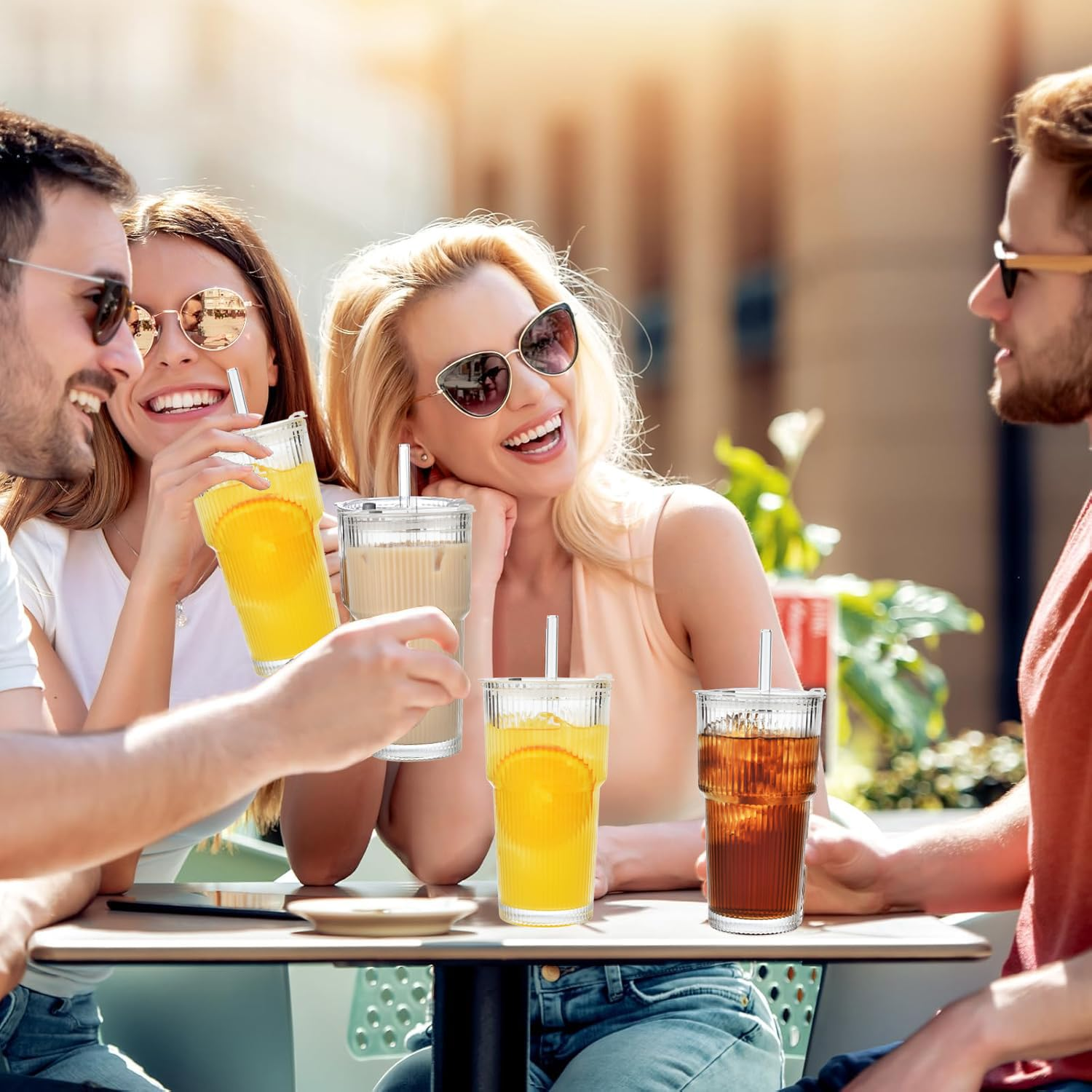 Vier Personen mit Sonnenbrillen lachen, trinken aus Gläsern mit Strohhalmen. Café im Freien.