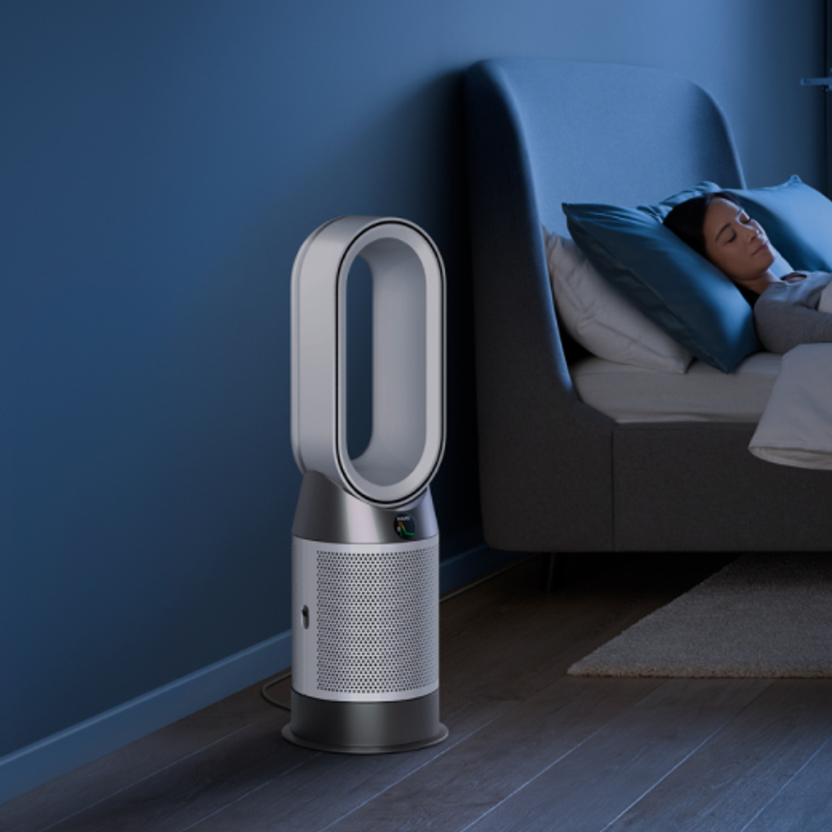 Una mujer duerme en la cama cerca de un ventilador Dyson. La habitación es oscura con paredes azules.