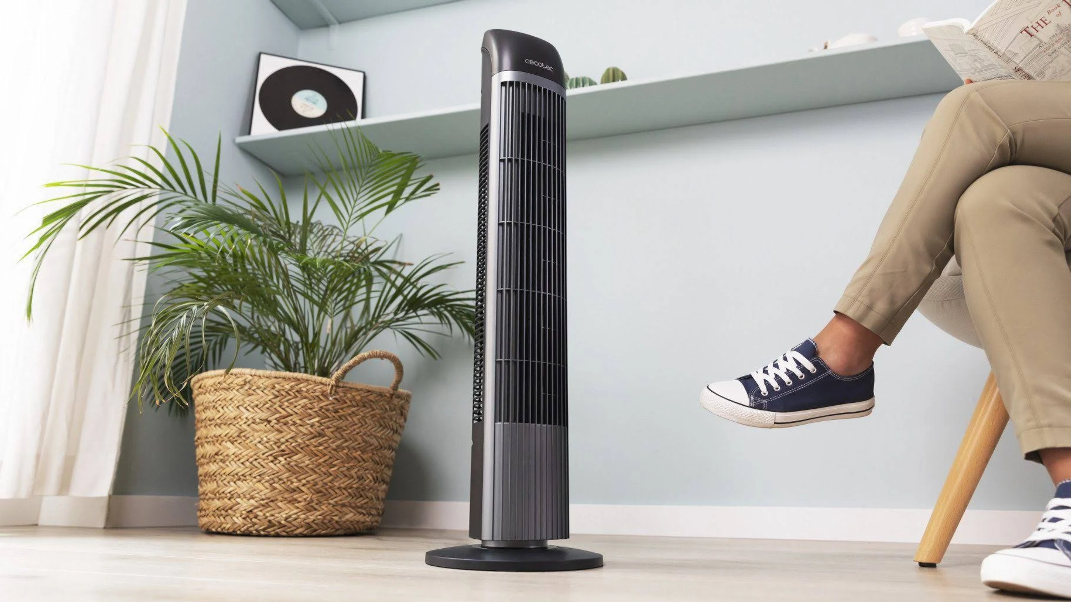 Wysoki, czarny wentylator wieżowy w pokoju, z osobą siedzącą w pobliżu, czytającą gazetę.