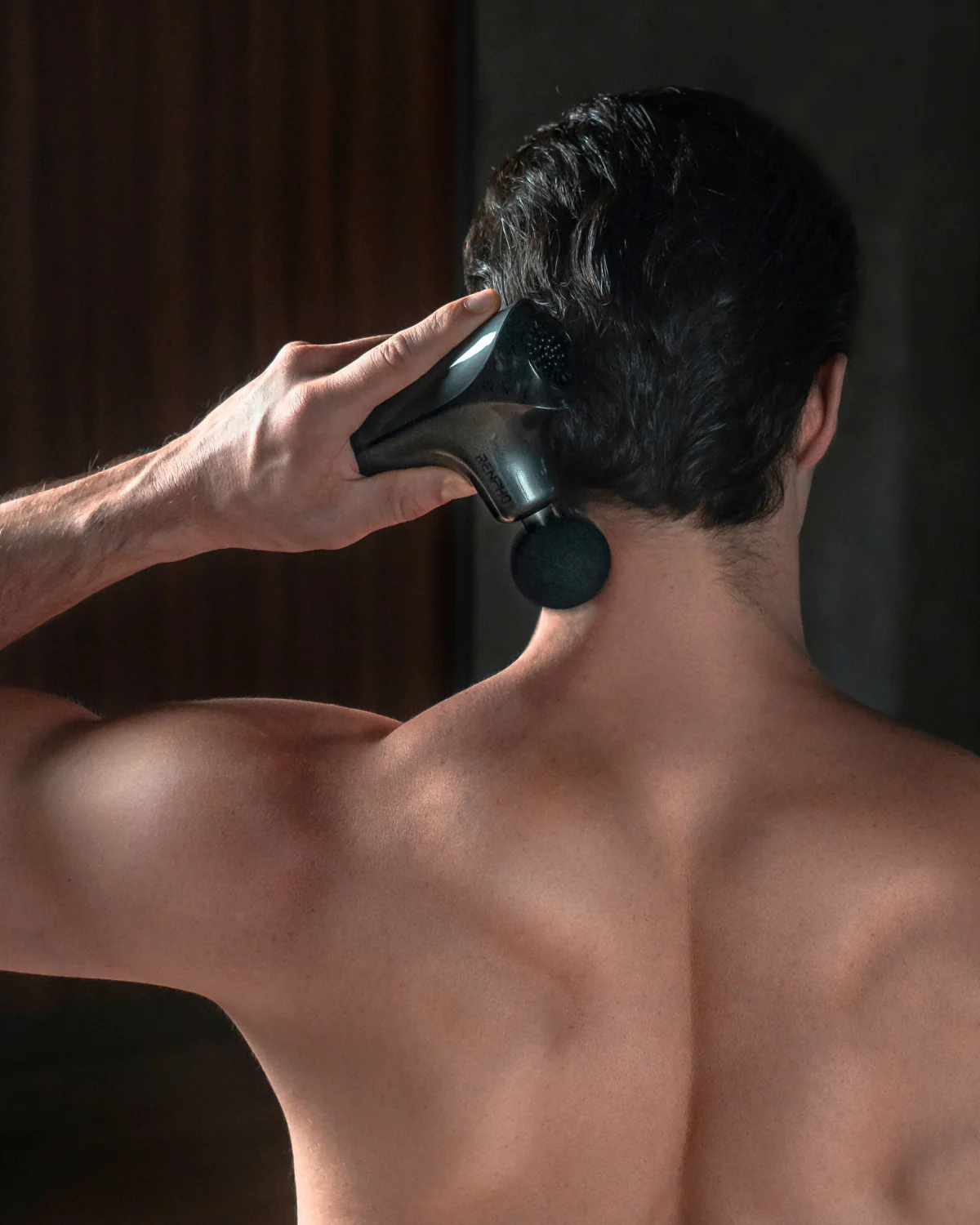 Una persona usando una pistola de masaje en el cuello, vista desde atrás, con enfoque en la pistola y la espalda.