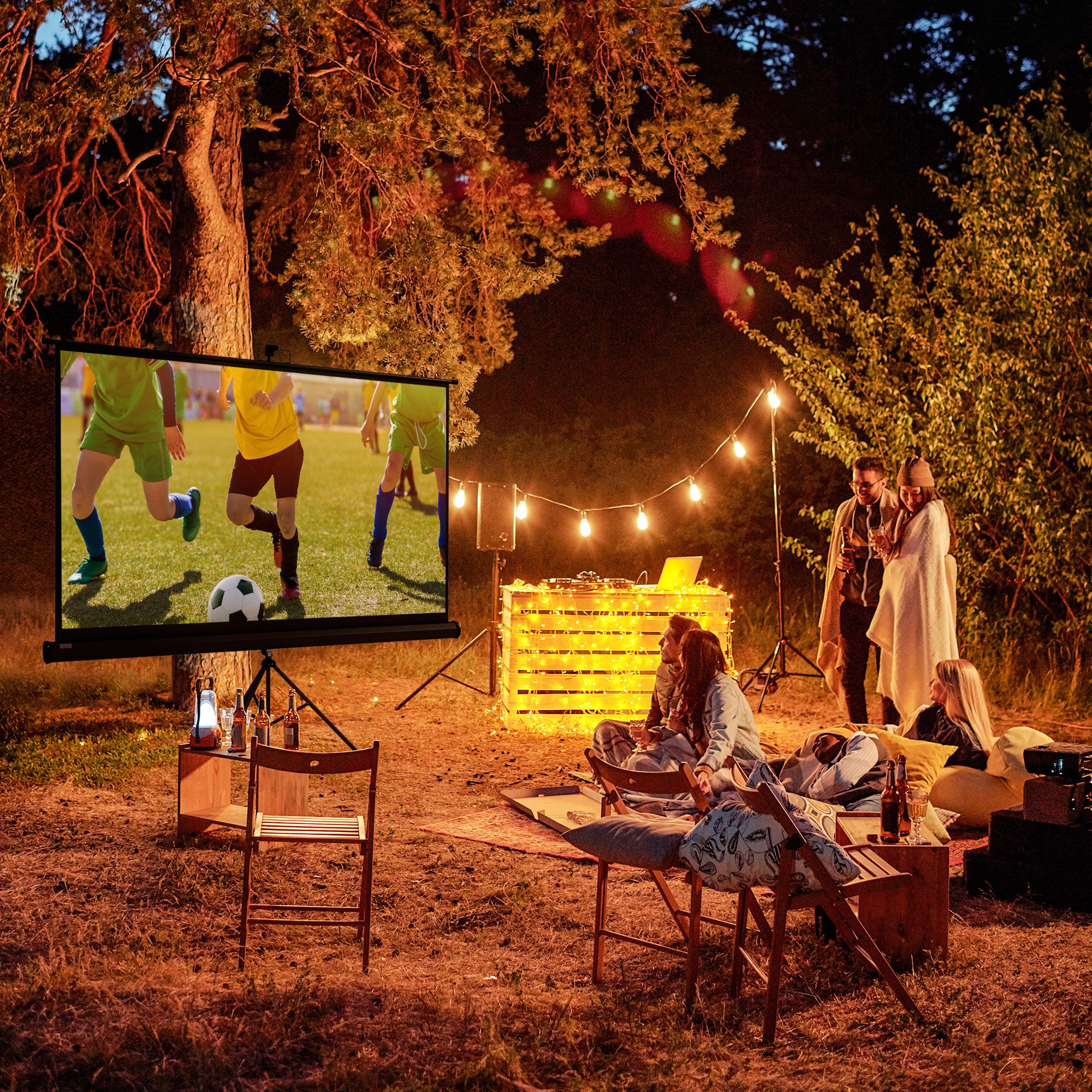 Uno schermo del proiettore mostra una partita di calcio. Le persone sono riunite intorno a un fuoco di notte.
