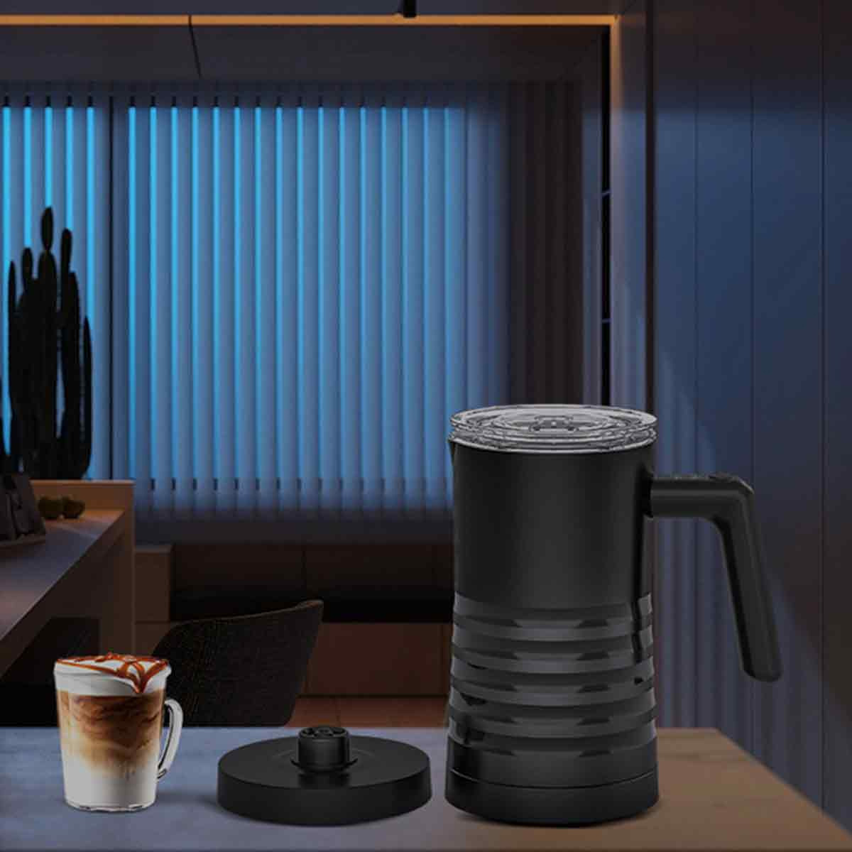 Ein schwarzer Milchaufschäumer auf einem Tisch. Ein Glas Latte Macchiato befindet sich links. Im Hintergrund sind Jalousien.