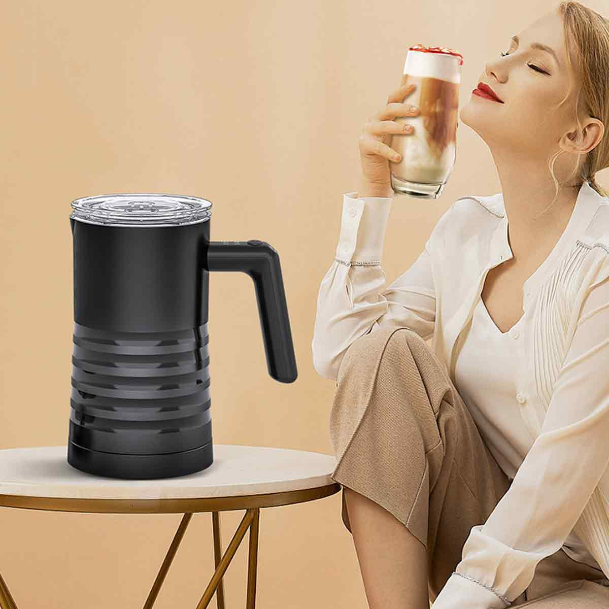 Frau hält ein Glas Latte Macchiato. Schwarzer Milchaufschäumer steht auf dem Tisch. Sie genießt das Getränk.