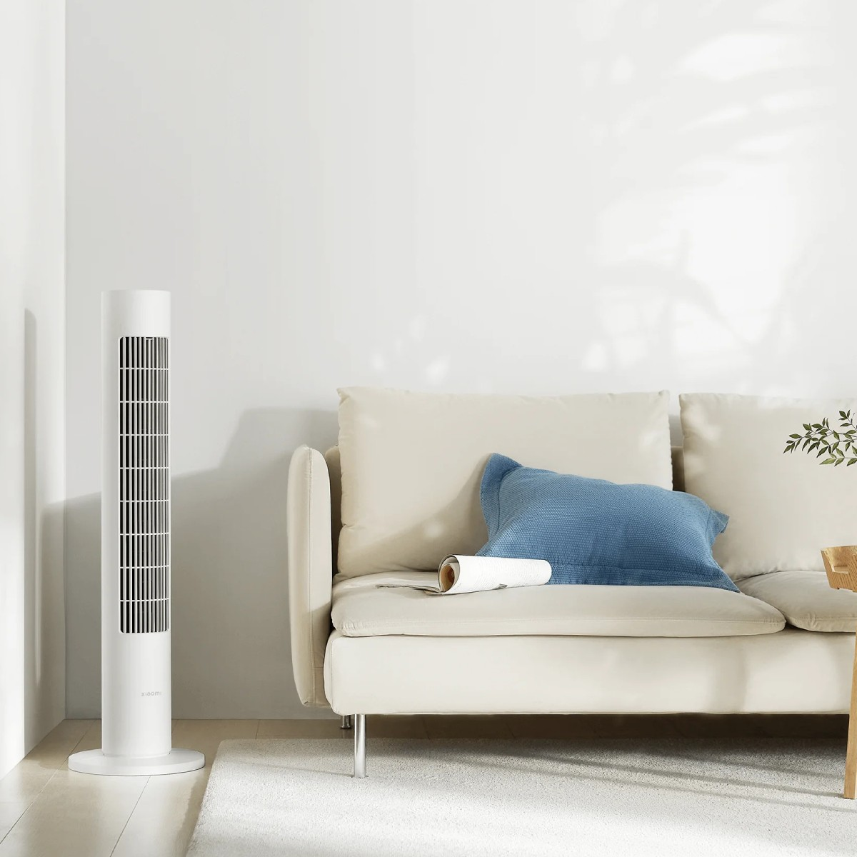Biały wentylator wieżowy obok beżowej sofy z niebieską poduszką i zwiniętym papierem.