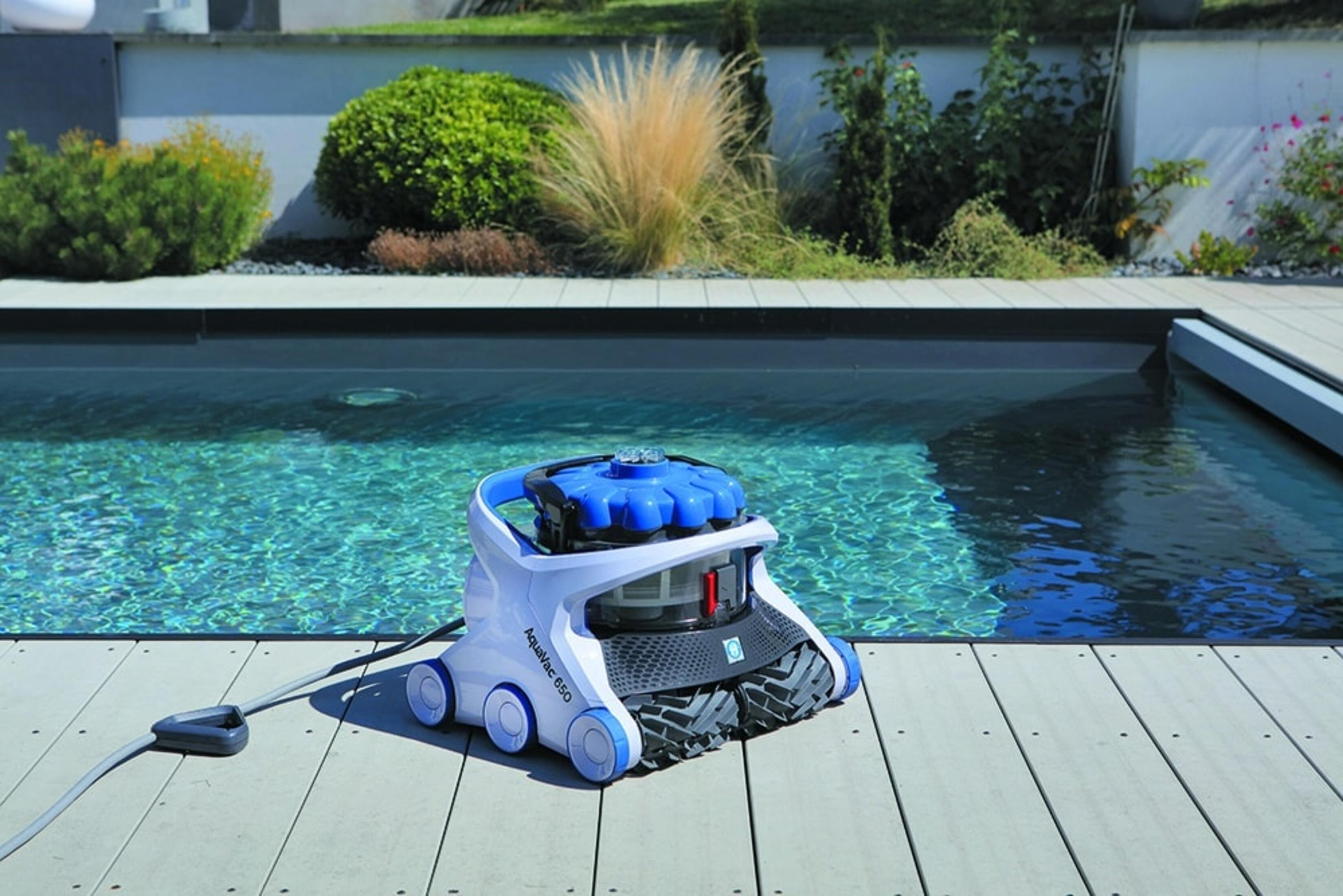 Robot limpiador de piscina en cubierta de madera junto a una piscina. El limpiador es blanco y azul.