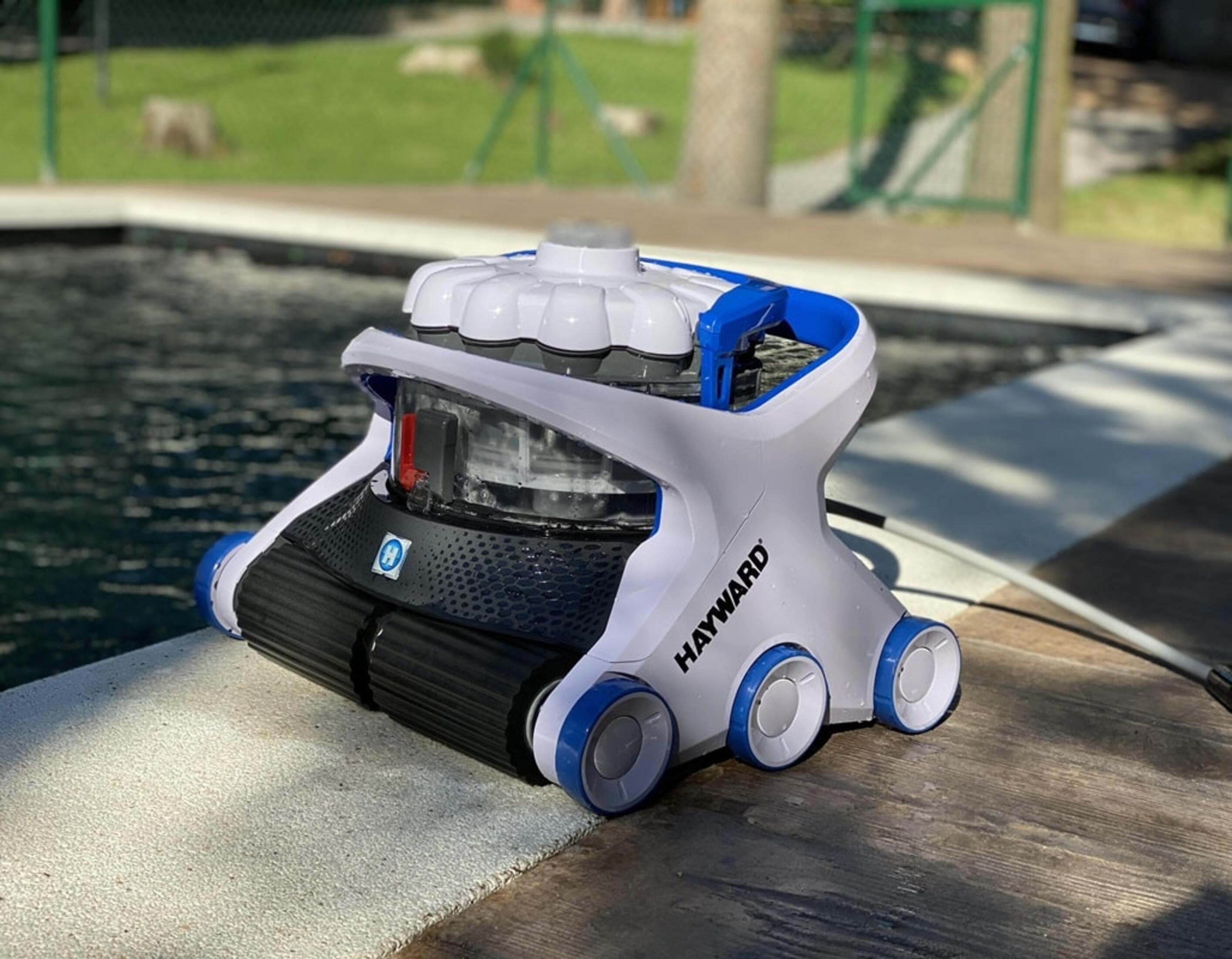 Un robot limpiador de piscinas blanco y azul al borde de una piscina, con un árbol al fondo.