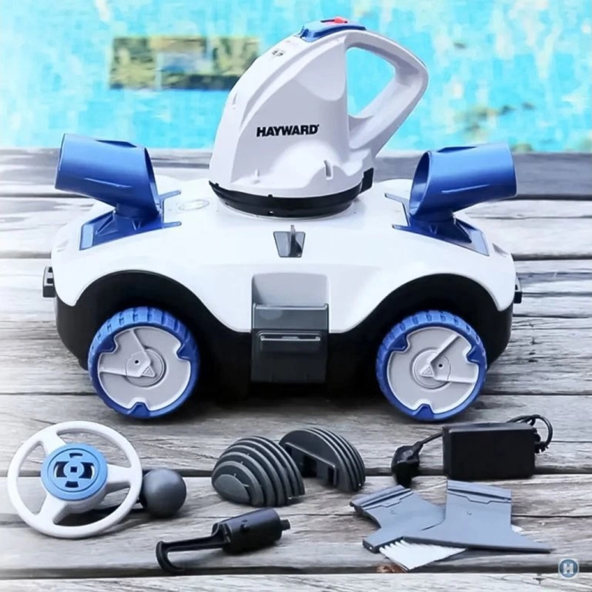 Limpiador de piscinas blanco y azul con accesorios, colocado sobre una superficie de madera junto a una piscina.