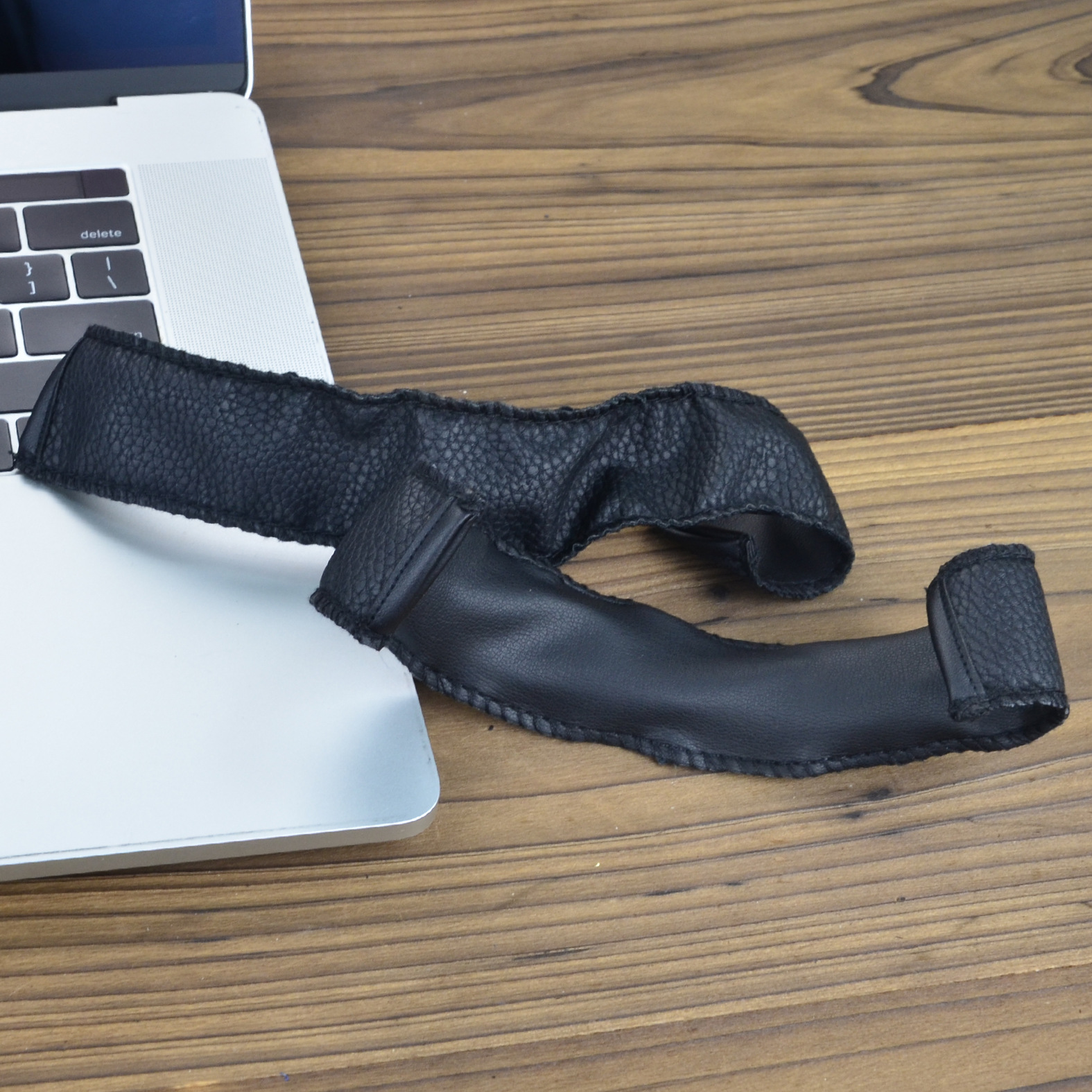 Una striscia di pelle nera appoggia su un laptop e un tavolo di legno. Il laptop è argentato e il tavolo è marrone scuro.