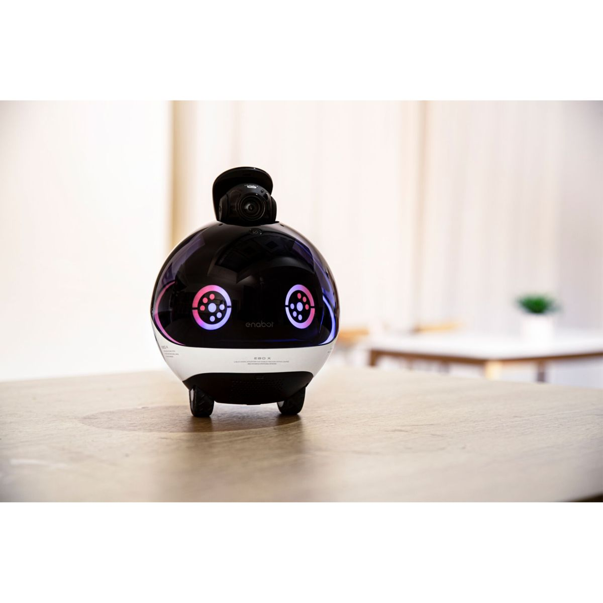 Un robot bianco e nero con una telecamera sopra. Ha luci colorate ed è su un tavolo di legno.