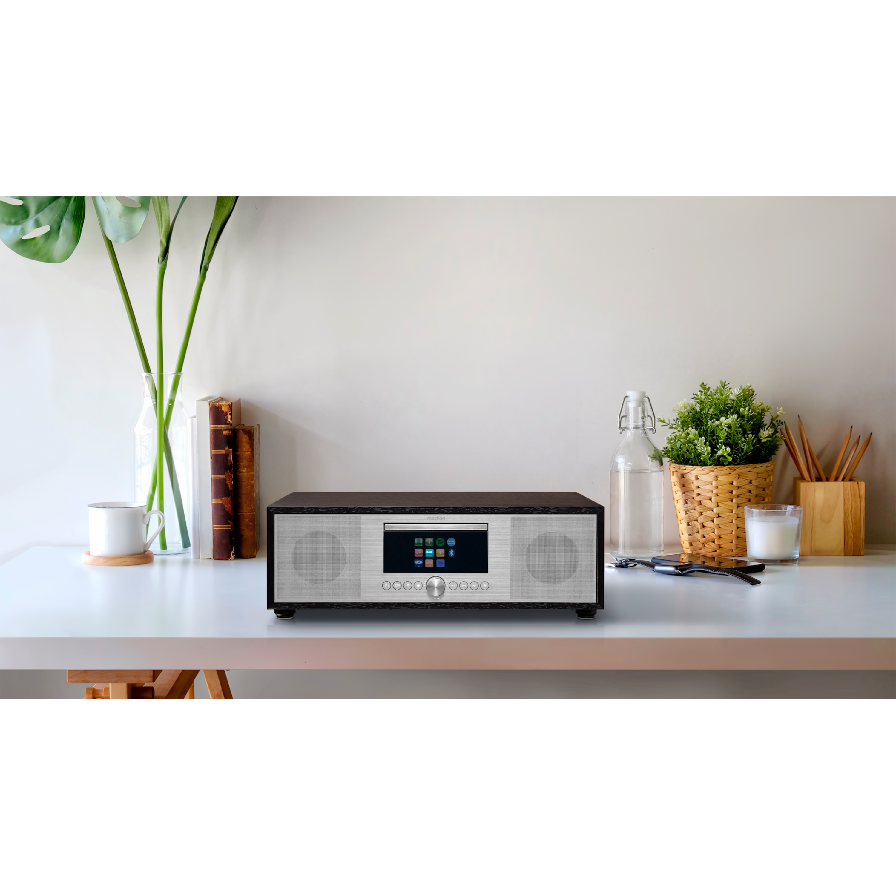 Un sistema de sonido negro y plateado sobre un escritorio blanco. Hay plantas y otros objetos en el fondo.