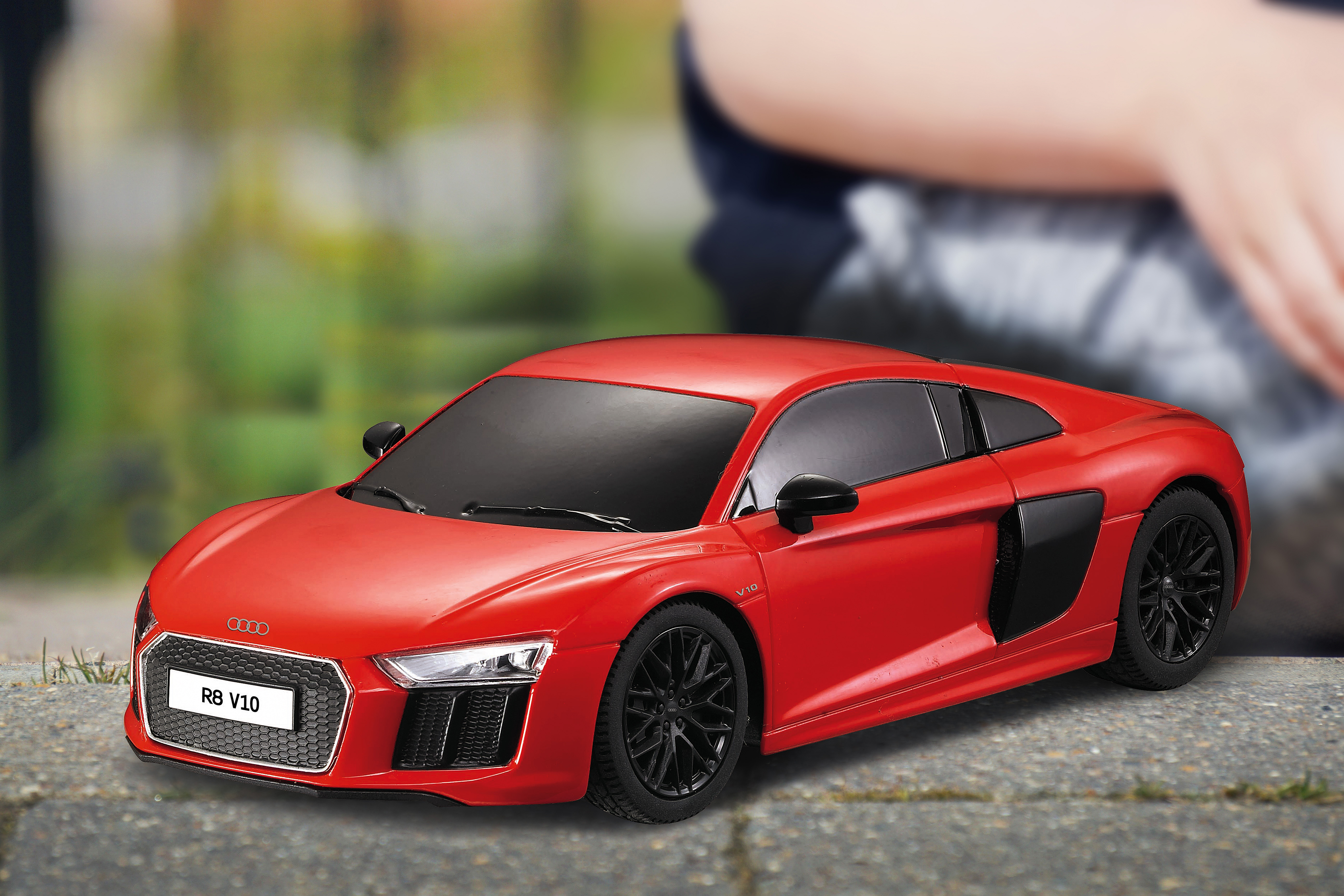 Un coche de juguete Audi R8 rojo sobre piedra, persona borrosa detrás.