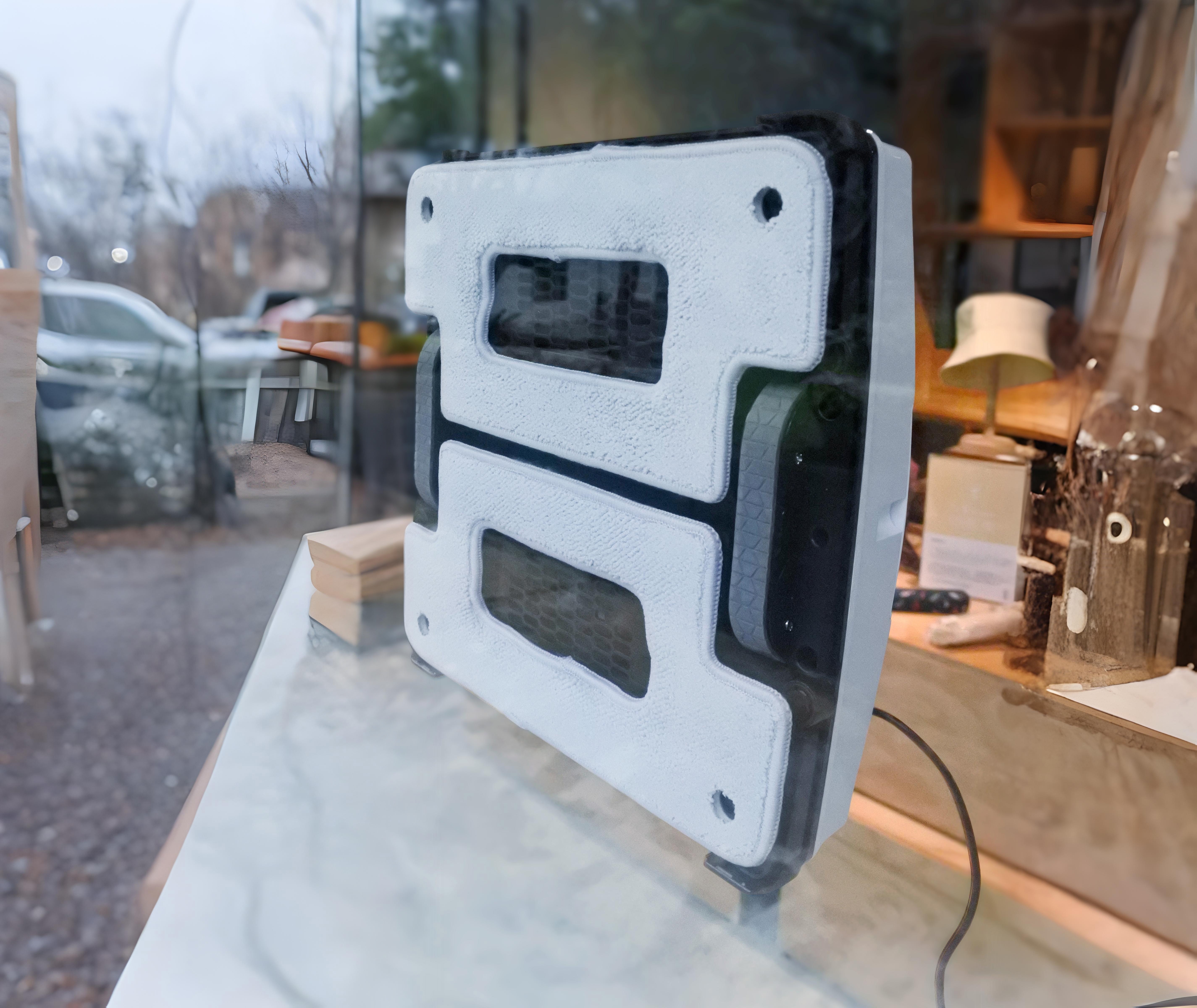 Se muestra un robot blanco de limpieza de ventanas detrás de una ventana de vidrio.