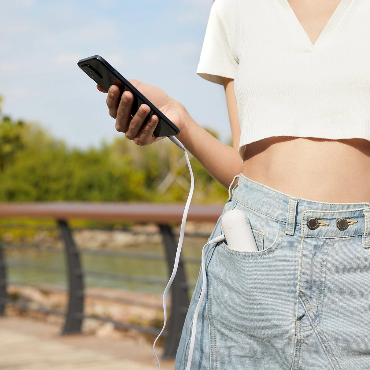 Persona che tiene un telefono nero collegato a un cavo bianco. Il cavo va in un piccolo dispositivo bianco in una tasca di jeans.