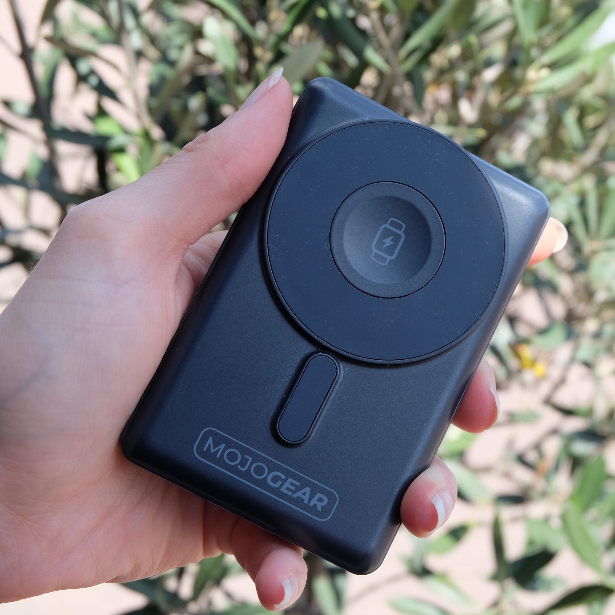 Een hand houdt een zwarte MOJOGEAR powerbank vast met een cirkelvormige oplaadpad tegen een groene achtergrond.