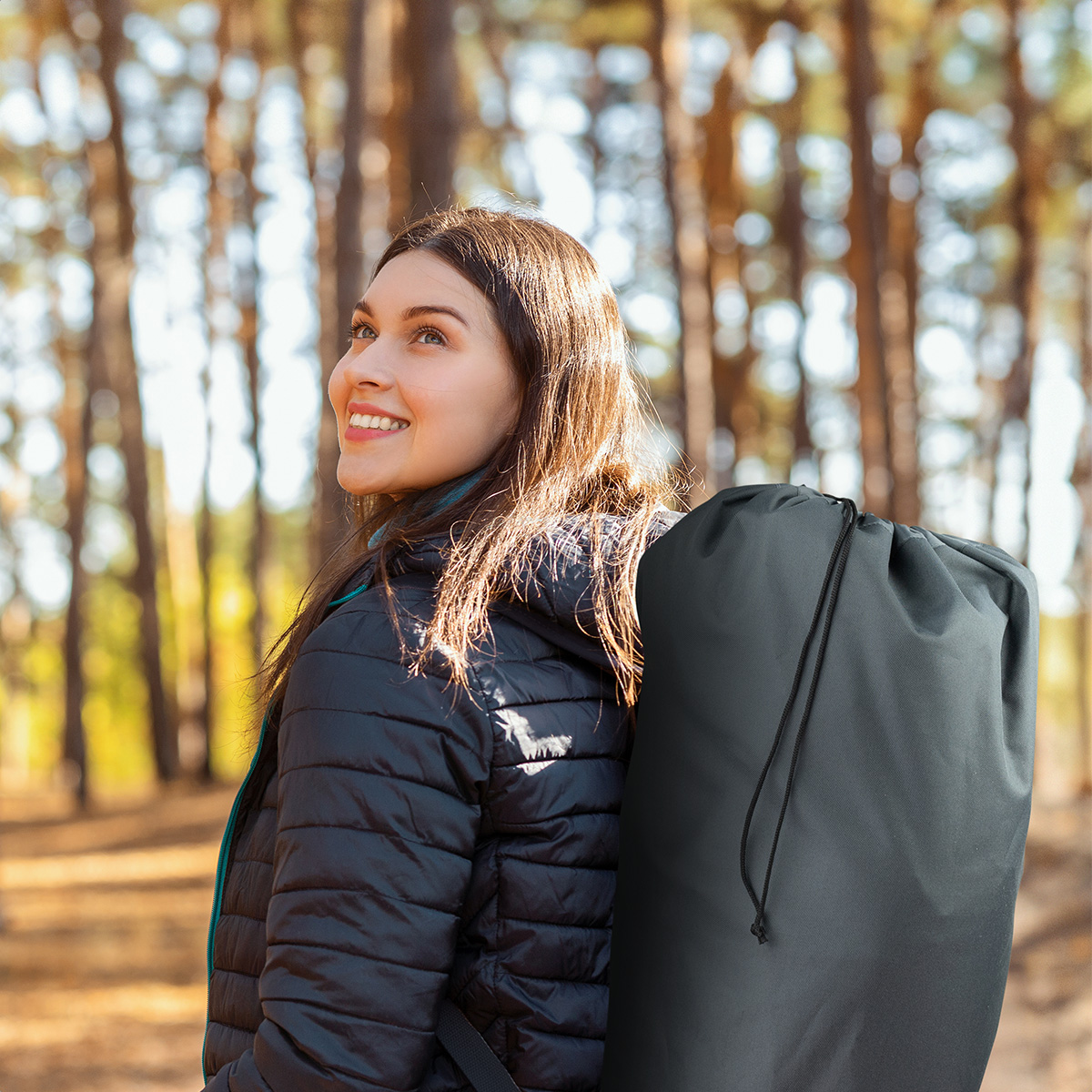 A woman is walking in a forest carrying a gray carrying bag over her shoulder. She smiles up at the trees.