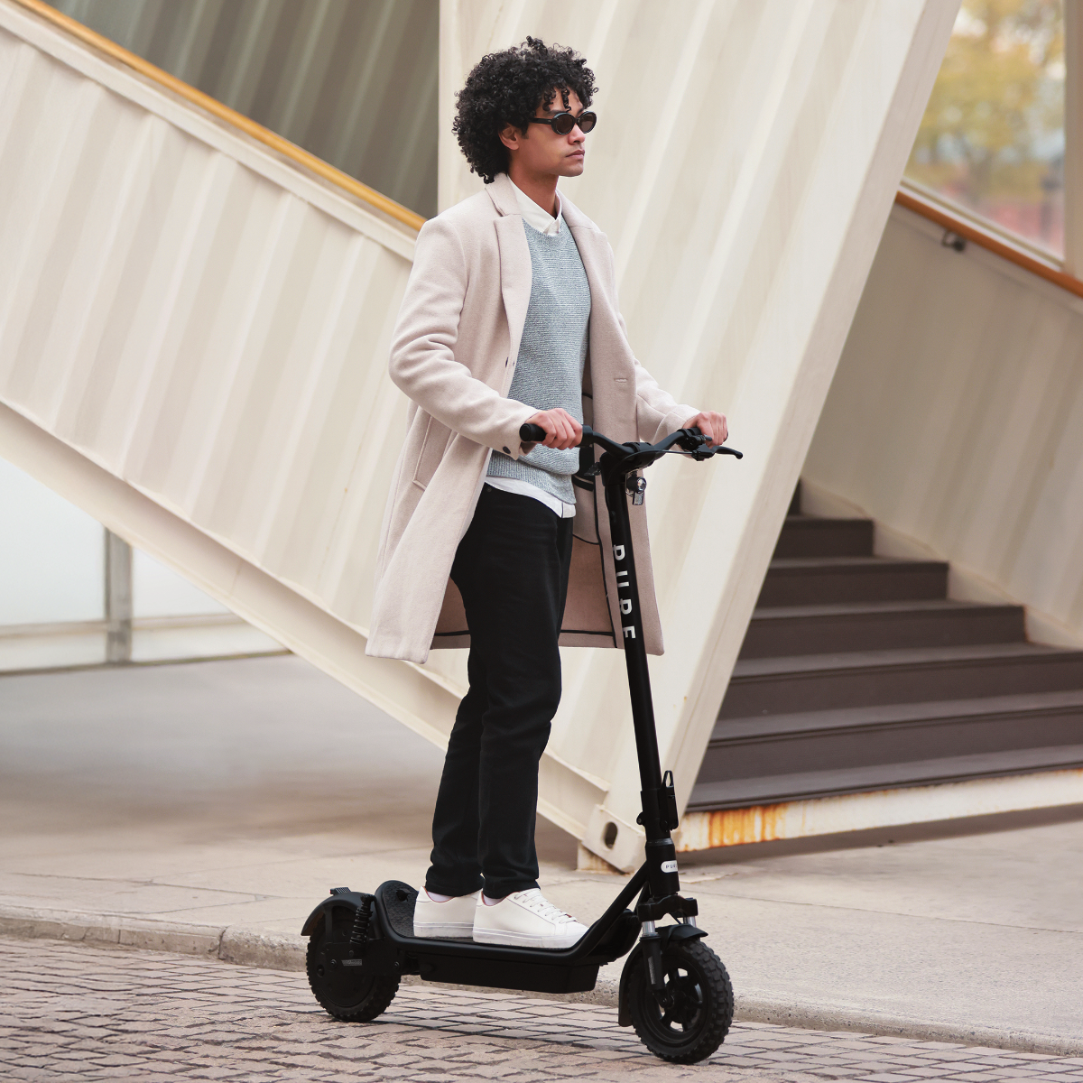 Mann auf einem Roller mit Sonnenbrille und Mantel. Er steht vor einem Gebäude mit Treppen.