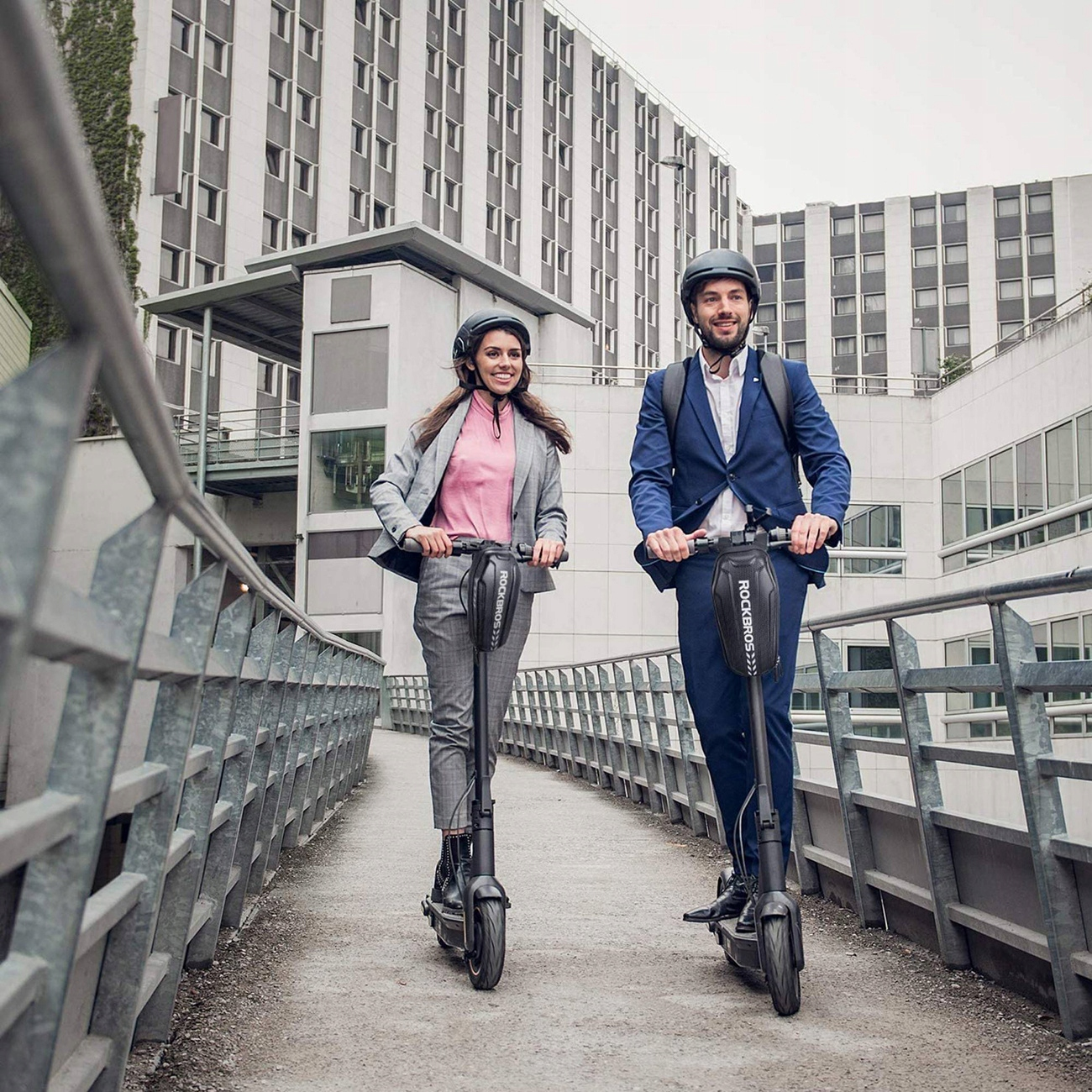 Dwie osoby na hulajnogach na moście. Mają kaski i stroje biznesowe. W tle budynki i barierka.